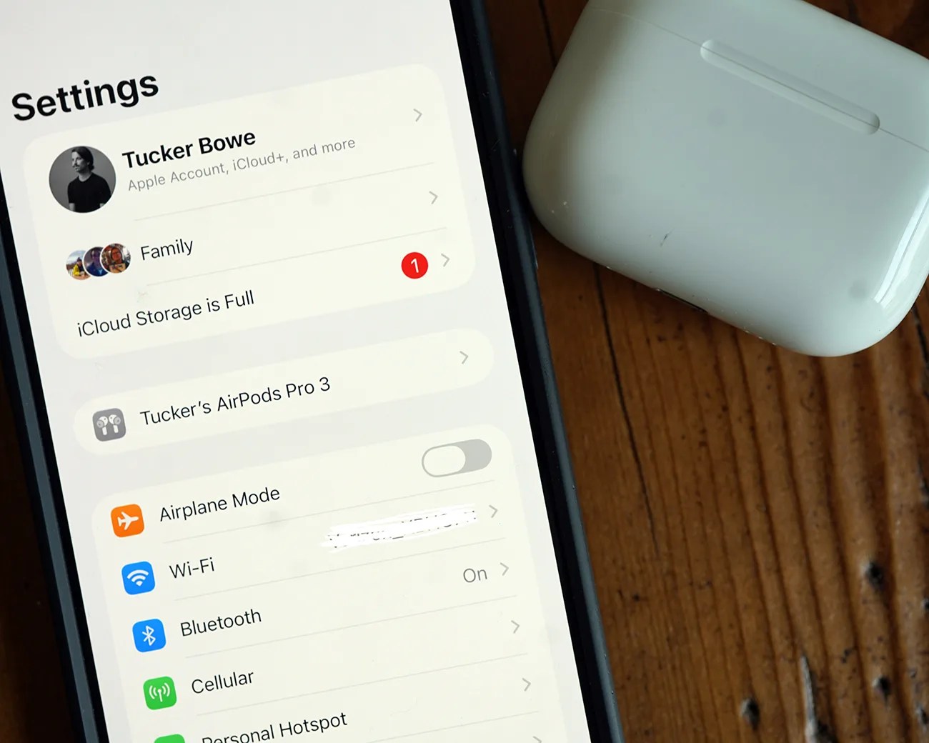 Smartphone screen showing iPhone settings menu with AirPods Pro 3 connected and white AirPods Pro charging case on wooden surface.