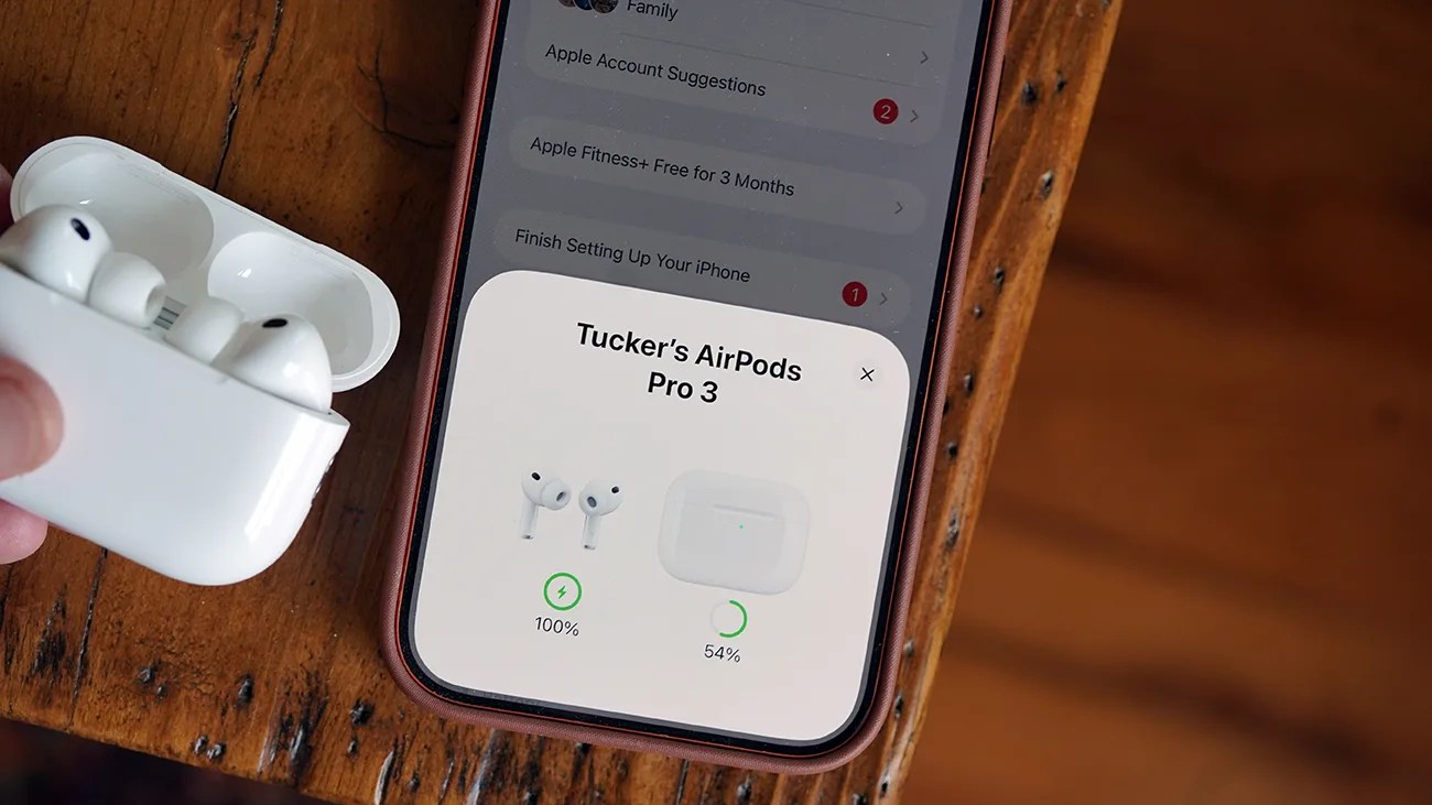 White AirPods Pro 3 earbuds in an open charging case held near a smartphone displaying their battery status.