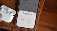 White AirPods Pro 3 earbuds in an open charging case held near a smartphone displaying their battery status.