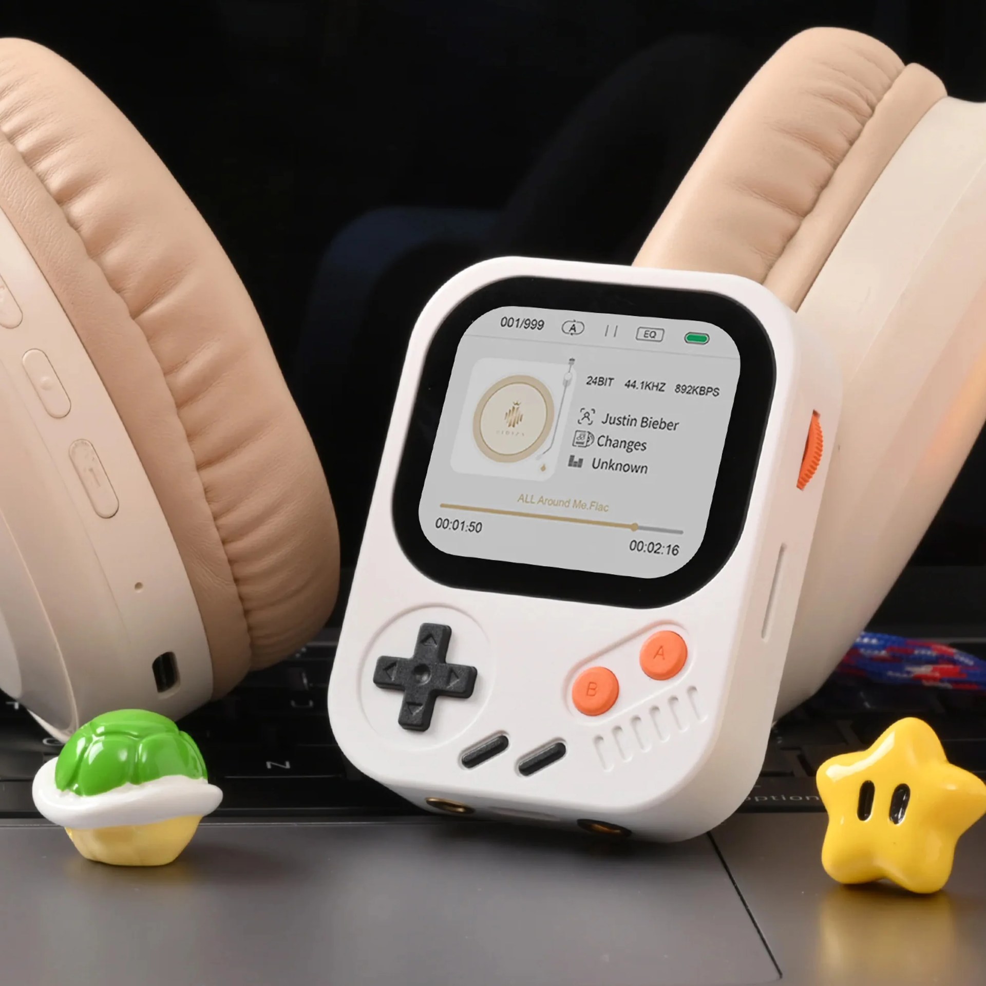White handheld gaming device with orange buttons and a screen showing a music player, placed between beige headphones on a keyboard.