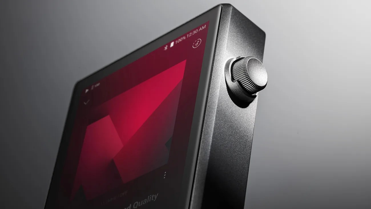 Close-up of a black digital audio player with a textured metal volume knob and a red geometric display screen.