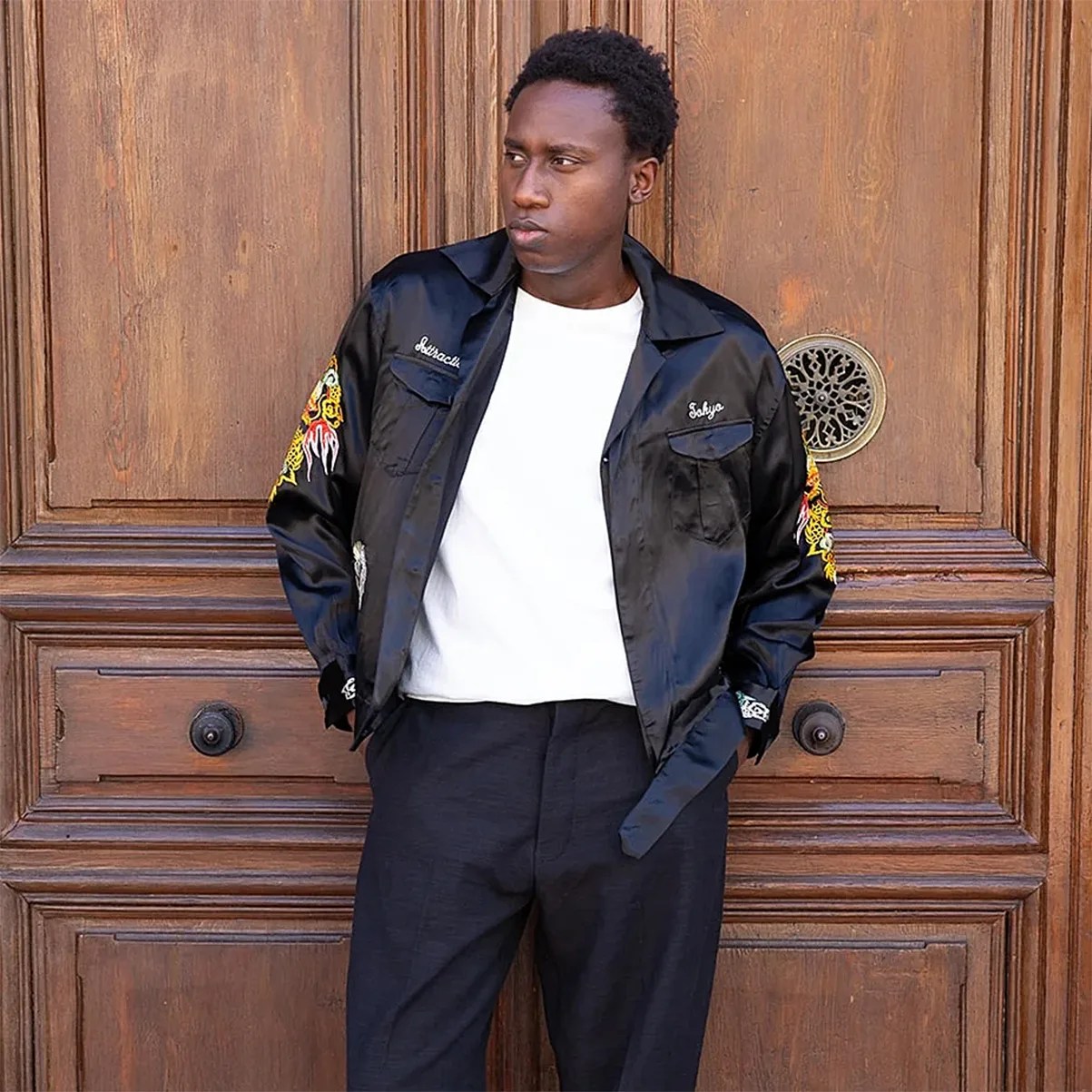 Man wearing a black satin jacket with embroidered yellow dragons on sleeves over a white shirt and black pants.