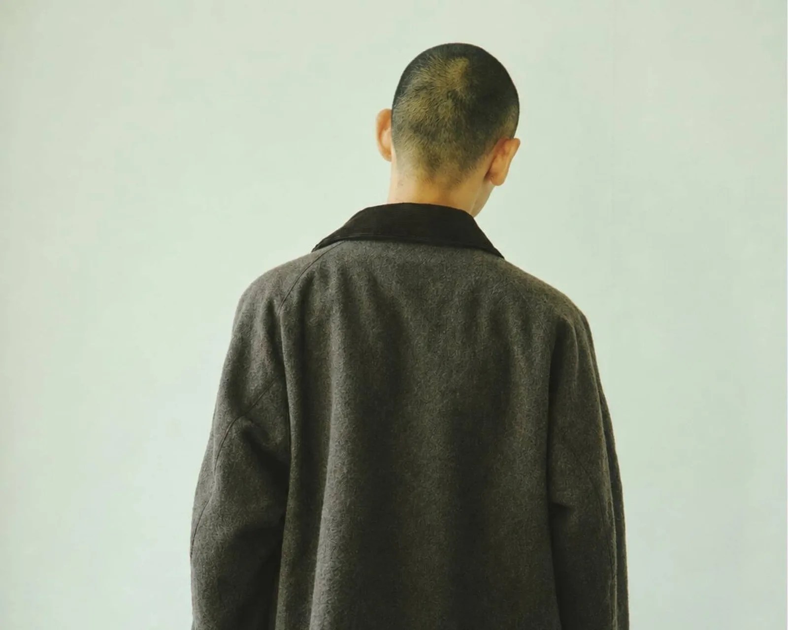 Person with short hair wearing a dark gray coat with a black collar, viewed from the back against a plain light background.