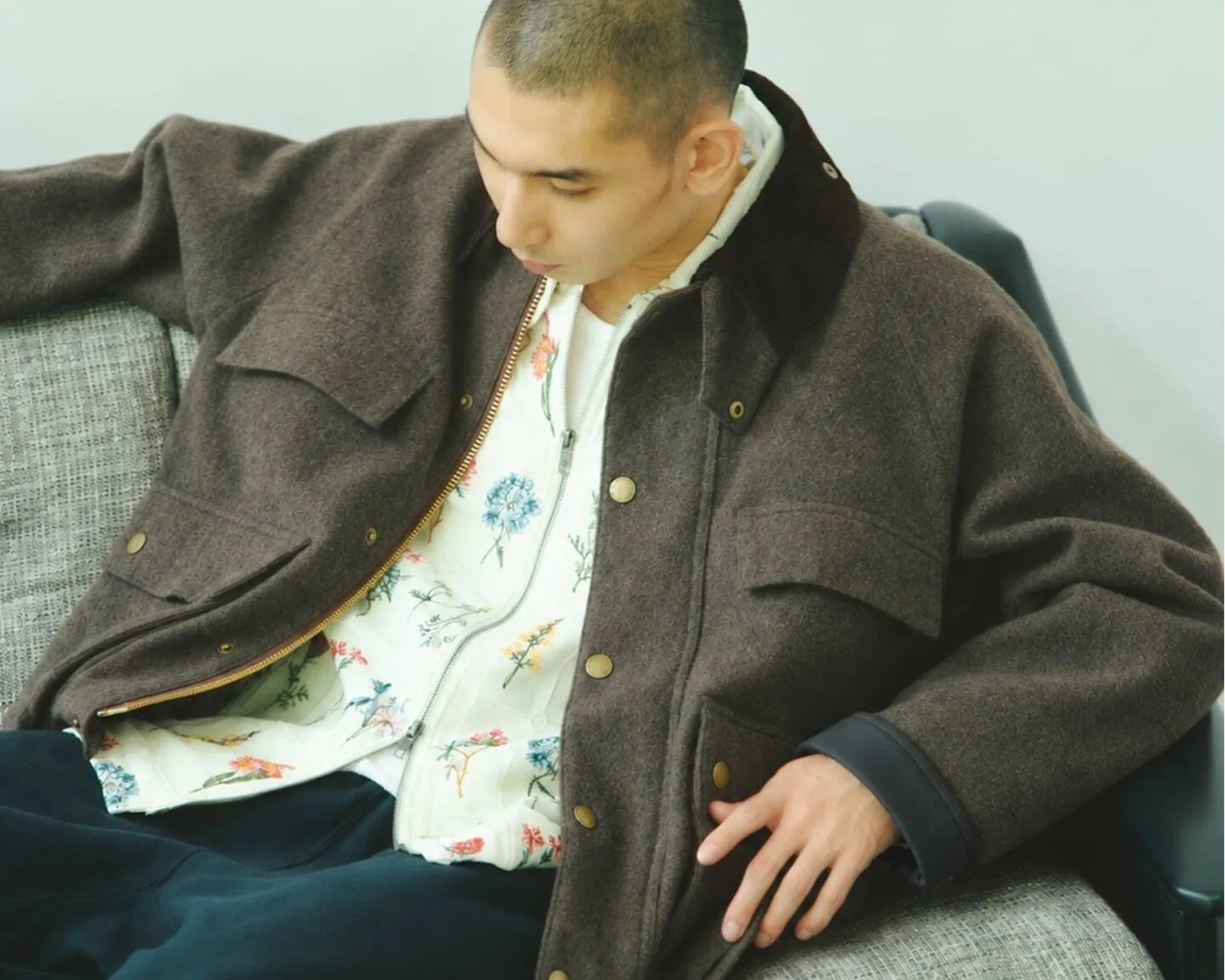 Man wearing a dark brown wool jacket over a white floral zip-up shirt sitting on a gray textured sofa.