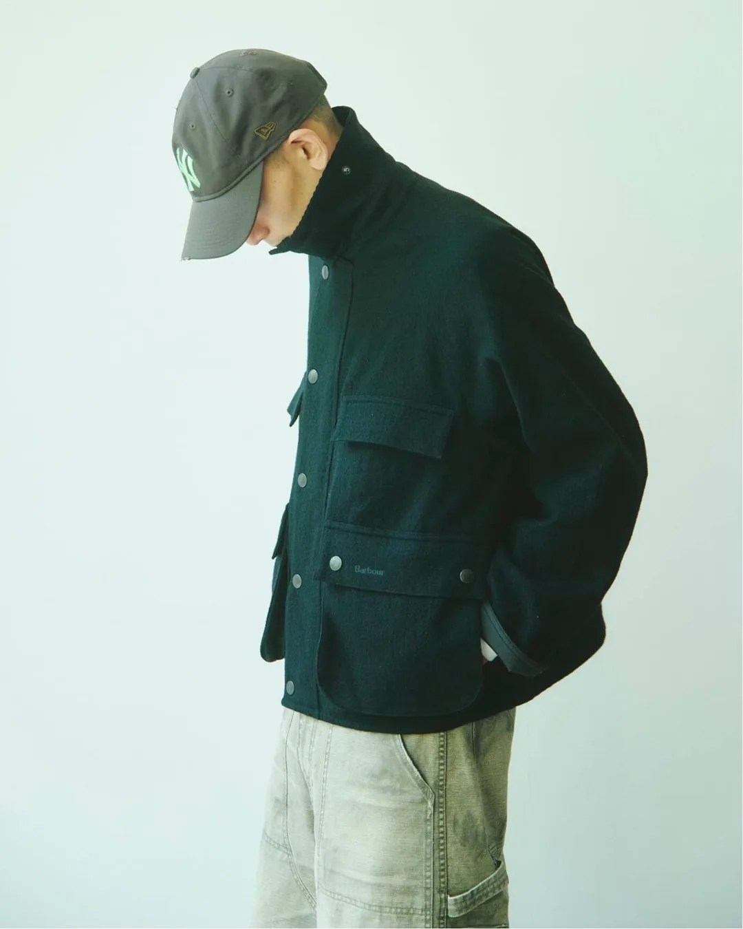 Man wearing a black button-up jacket with multiple pockets and light-colored pants, with a dark gray baseball cap.