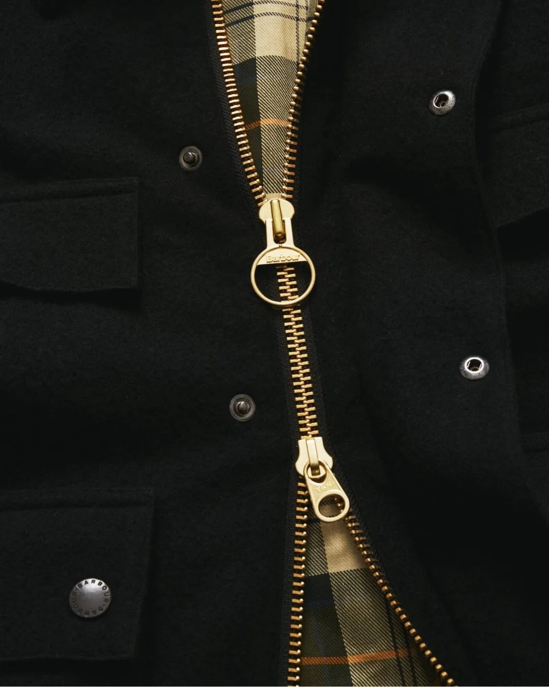 Close-up of a black jacket with gold zipper and snap buttons, showing a beige and green plaid lining.