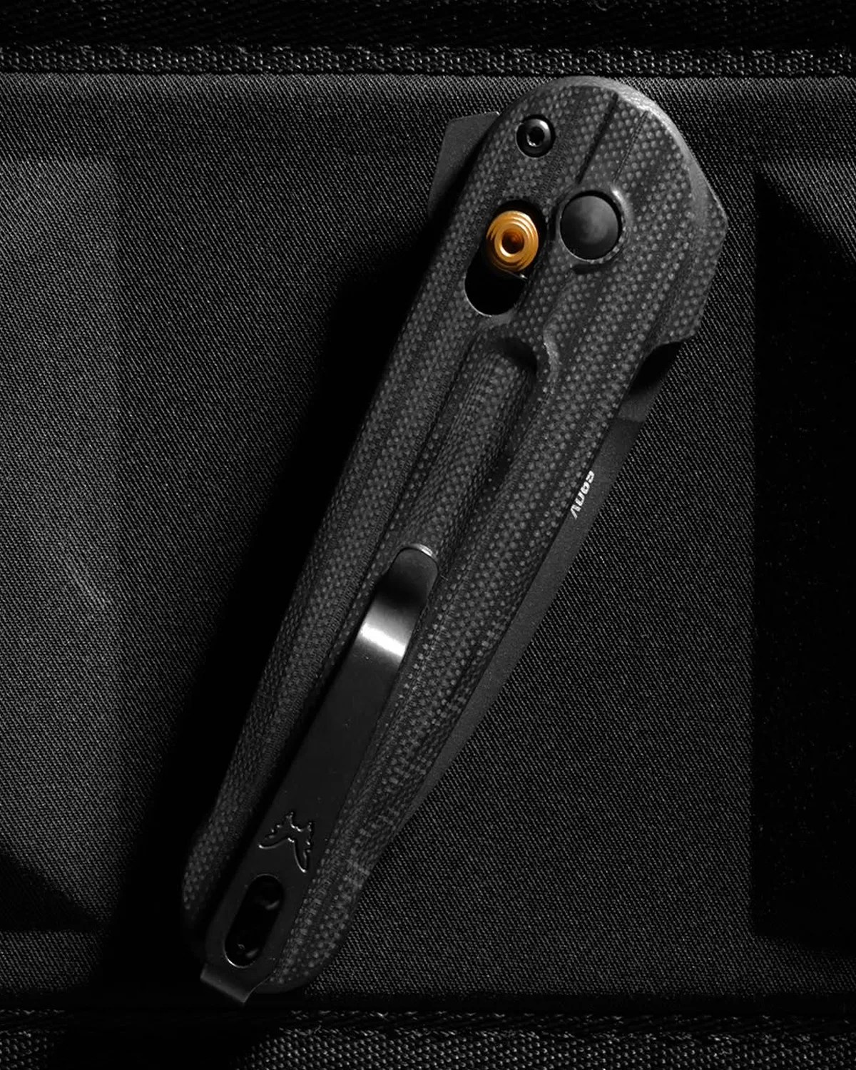 Black textured folding knife with a metal clip and a visible brass pivot screw on a black fabric surface.