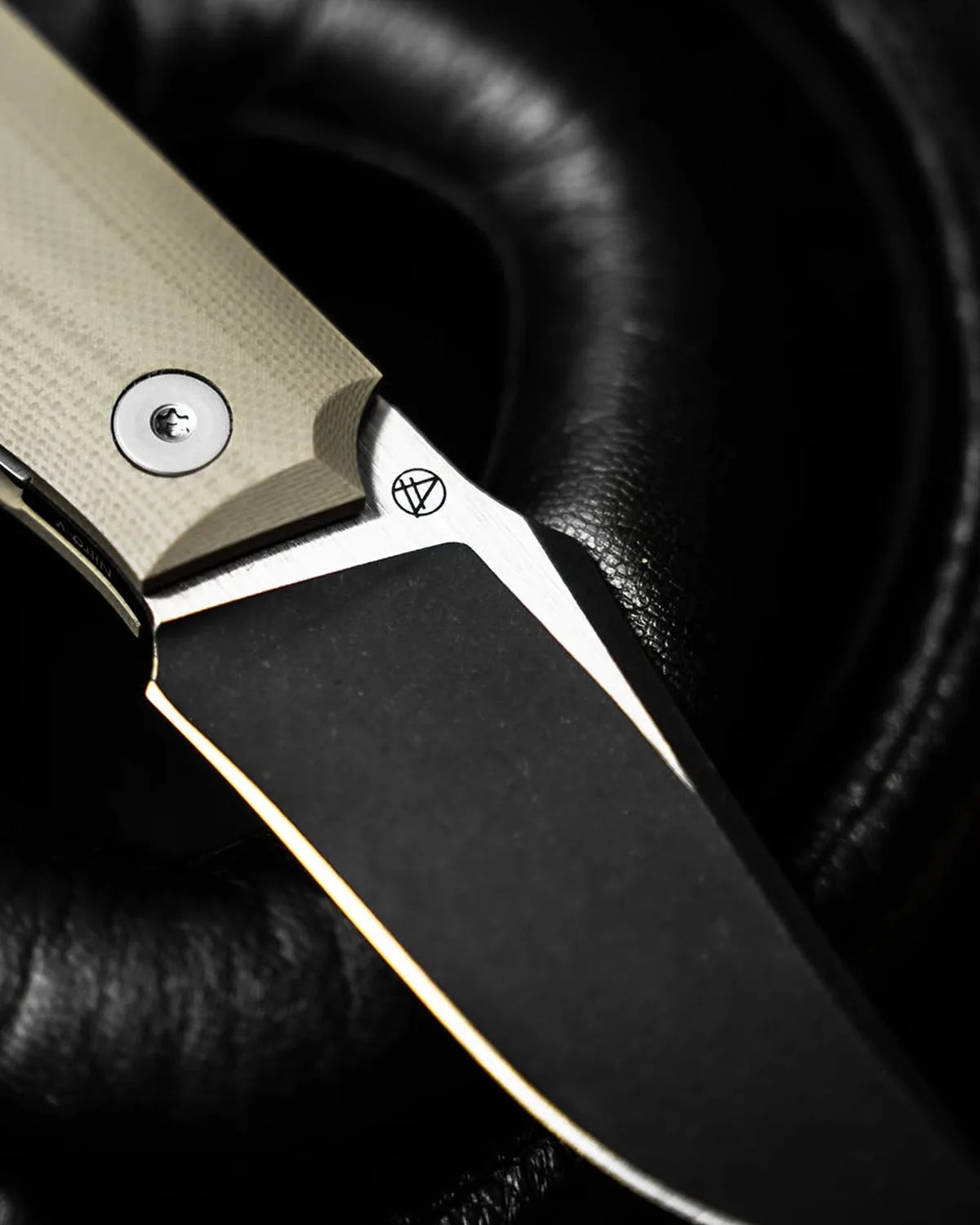 Close-up of a knife blade with a black and silver finish and a beige textured handle resting on black leather.