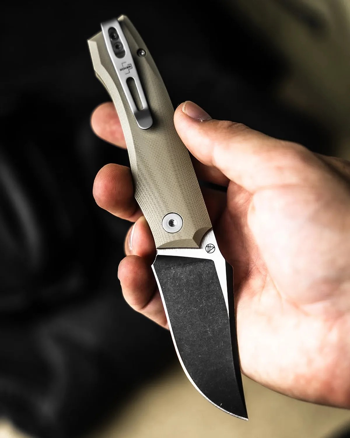 Hand holding a folding knife with a tan textured handle and a black stonewashed blade.