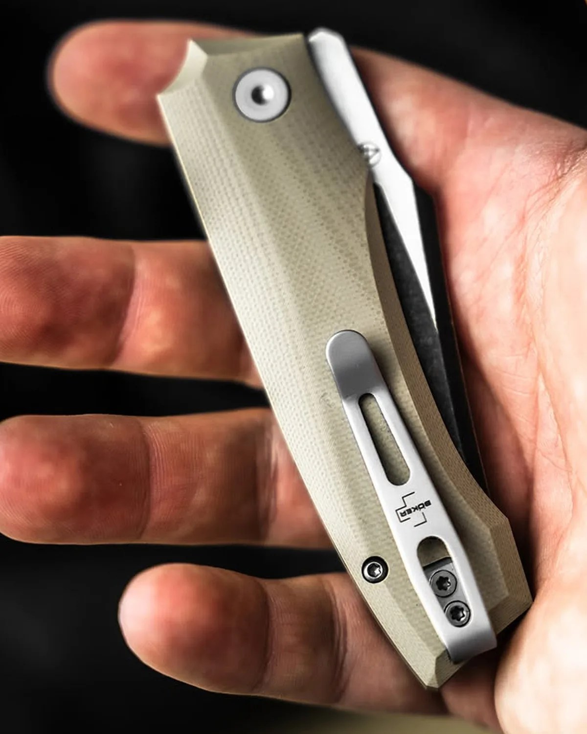 Beige textured folding knife with silver clip held in a hand against a black background.