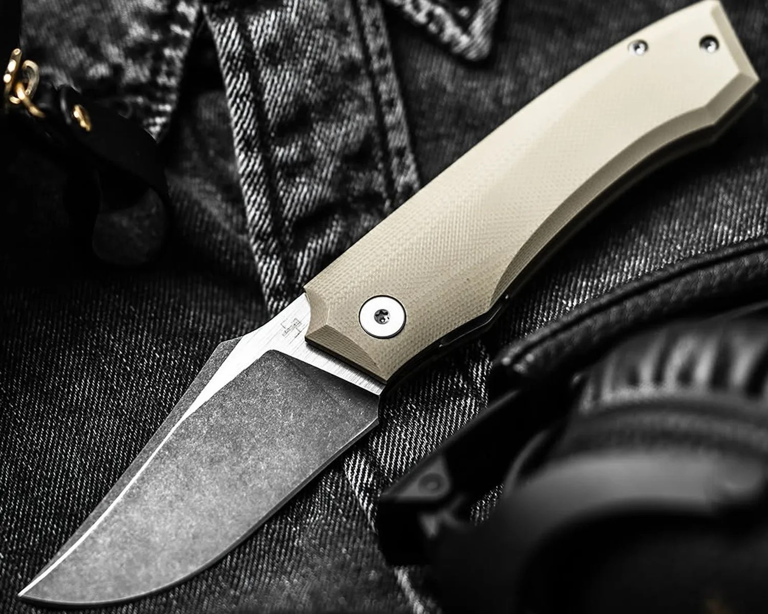Folding knife with a stonewashed blade and beige textured handle on dark denim fabric.
