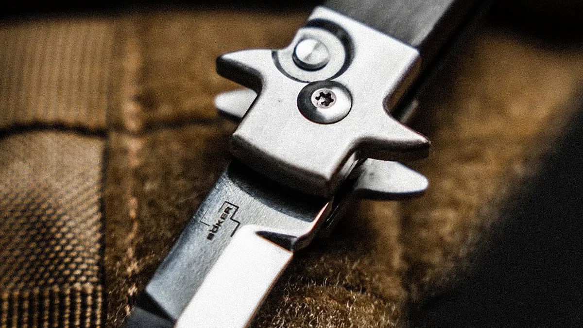Close-up of a silver folding knife blade with a partially visible "Boker" logo on a textured brown fabric surface.