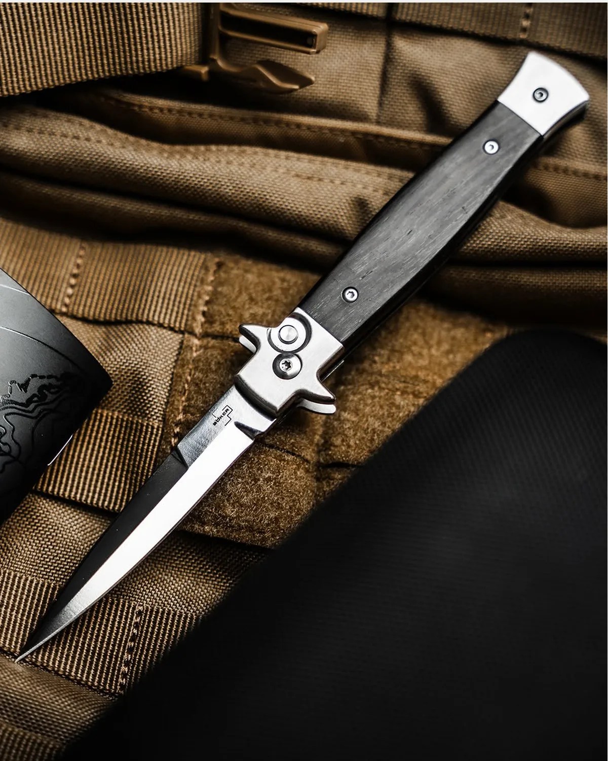 Folding knife with a black handle and silver bolsters resting on brown tactical fabric.
