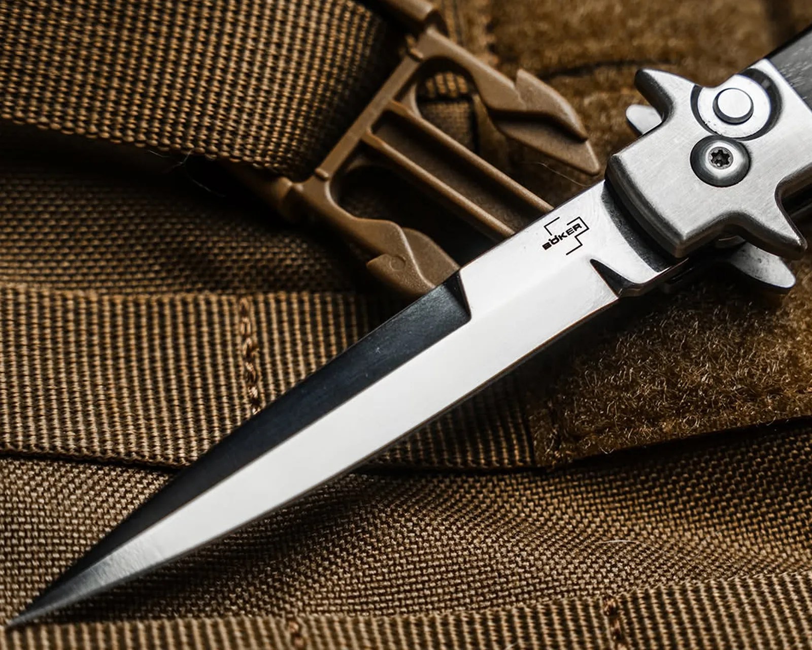 Close-up of a Böker folding knife blade with a sharp point resting on brown tactical fabric and strap.