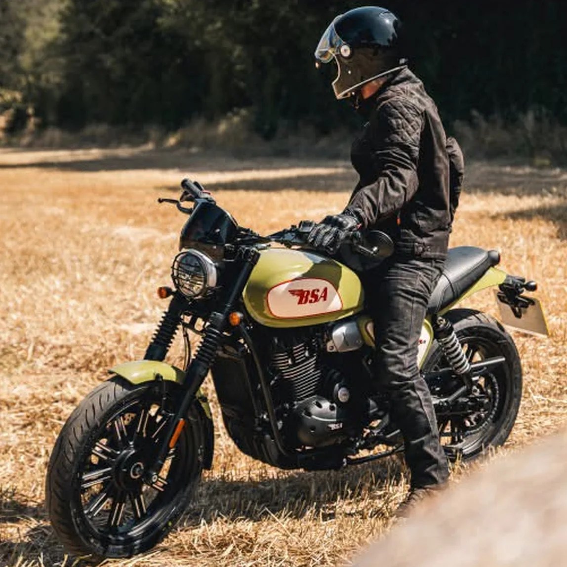 Person wearing black riding gear and helmet sitting on a green and black BSA motorcycle in a dry grassy field.