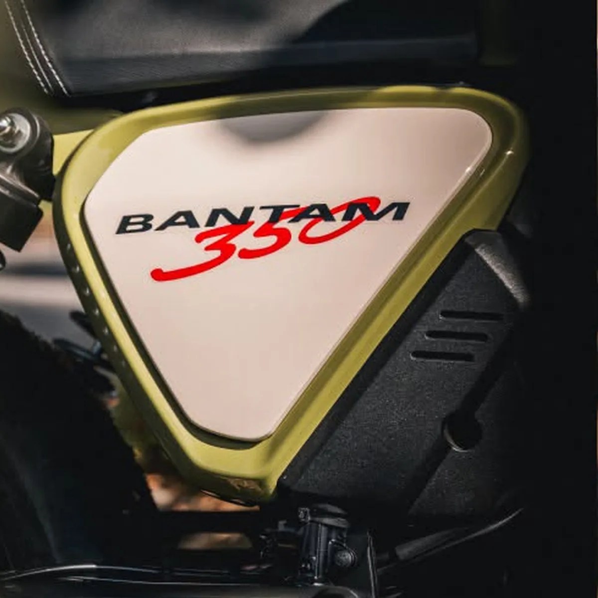 Close-up of a motorcycle side panel with "BANTAM 350" text in black and red on a cream background with olive green trim.