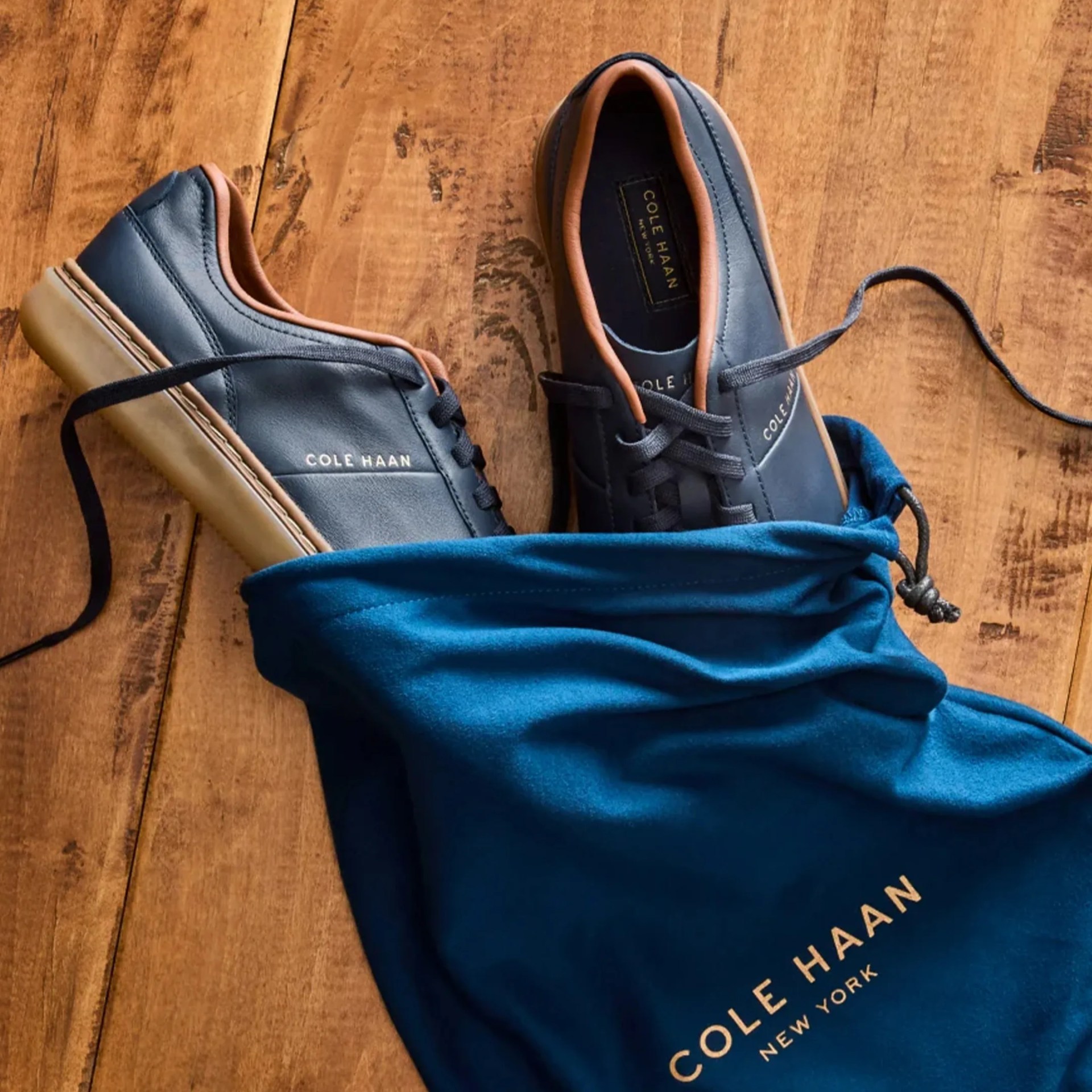 Pair of navy blue Cole Haan leather sneakers with tan trim and gum soles partially inside a blue Cole Haan drawstring bag on a wooden surface.