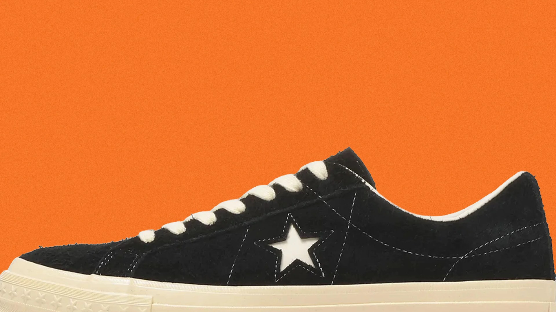 Black suede sneaker with white star logo and white laces on an orange background.