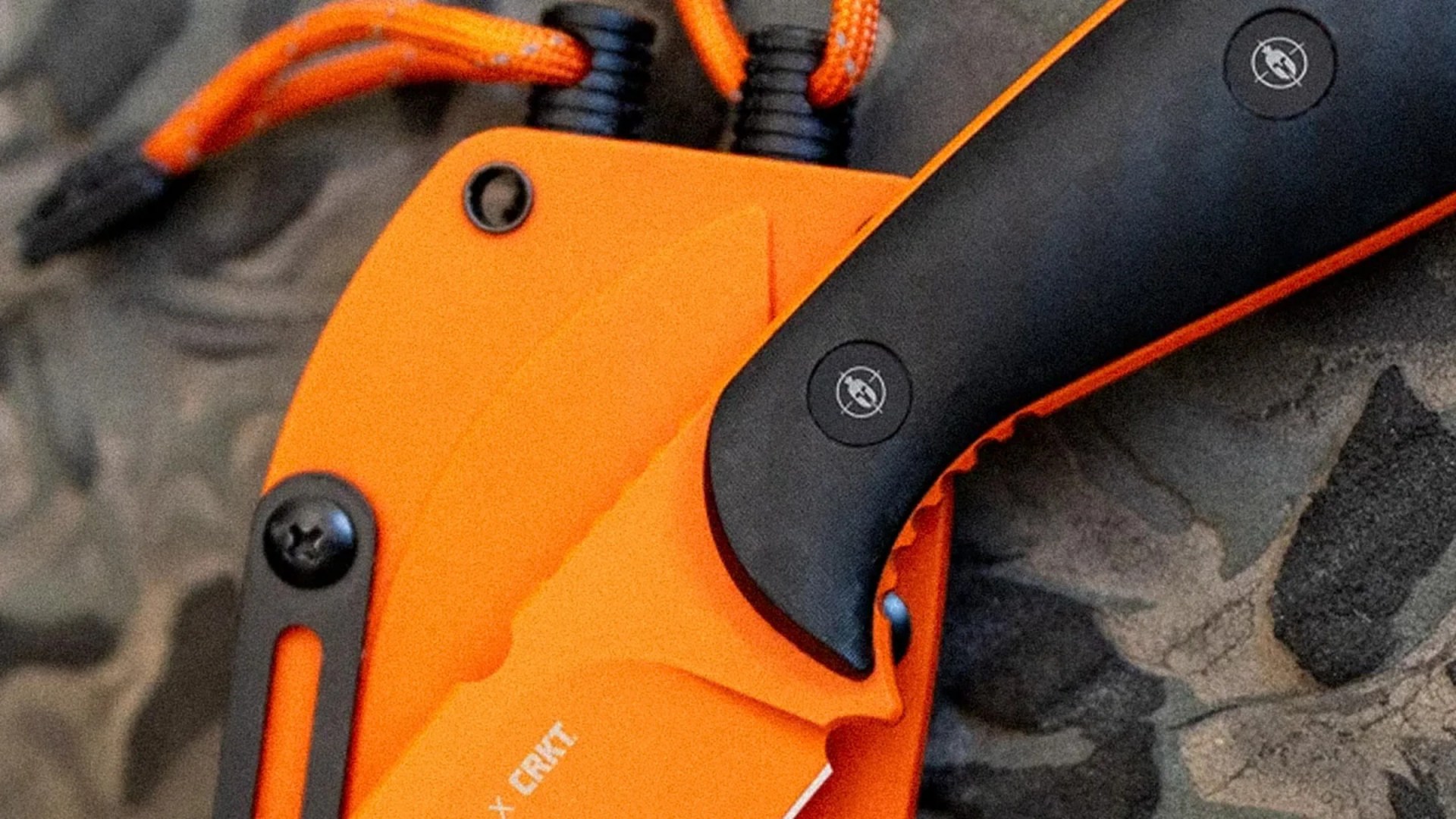 Close-up of an orange CRKT knife sheath with black handle and orange paracord on camouflage fabric.