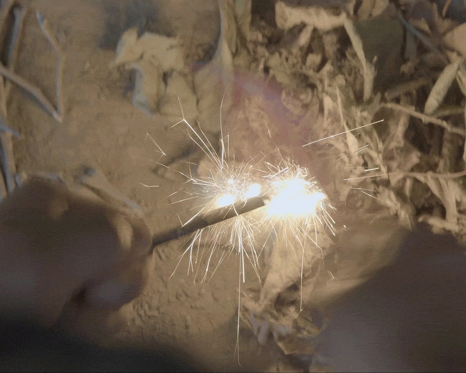 Two sparklers burning with bright sparks held close together over a dirt and dry leaves ground.