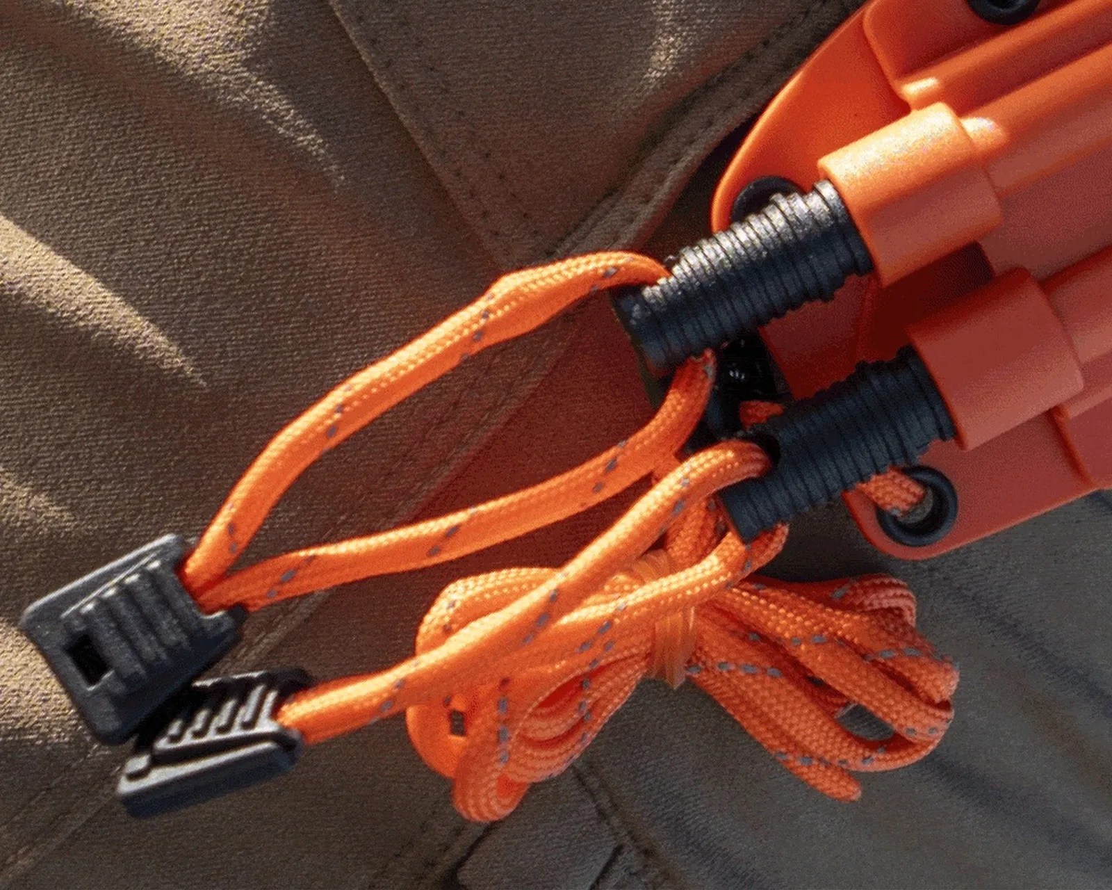 Close-up of orange bungee cords with black plastic hooks attached to an orange plastic device on a brown fabric surface.