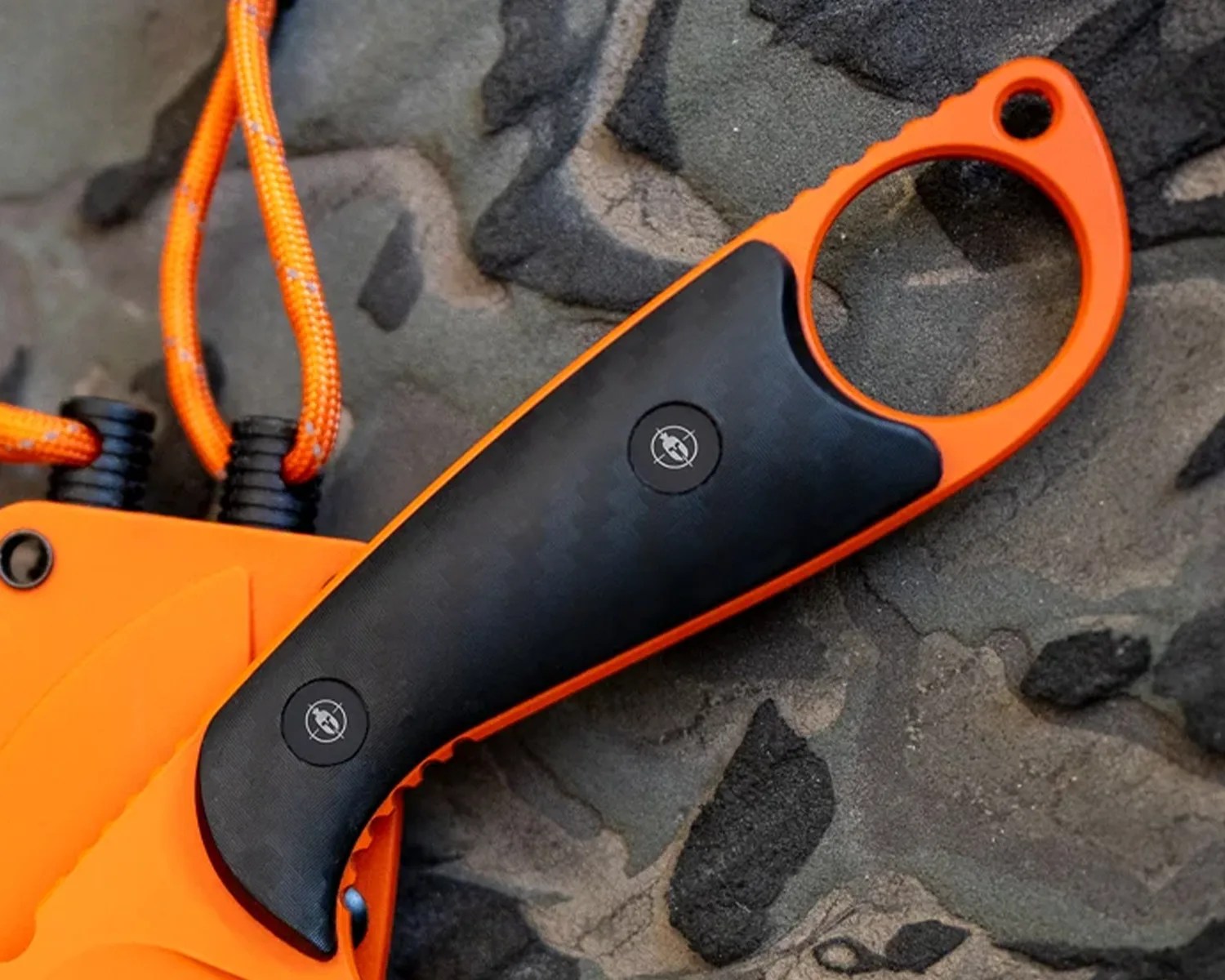 Orange and black knife handle with a finger hole and attached orange cord on a textured surface.