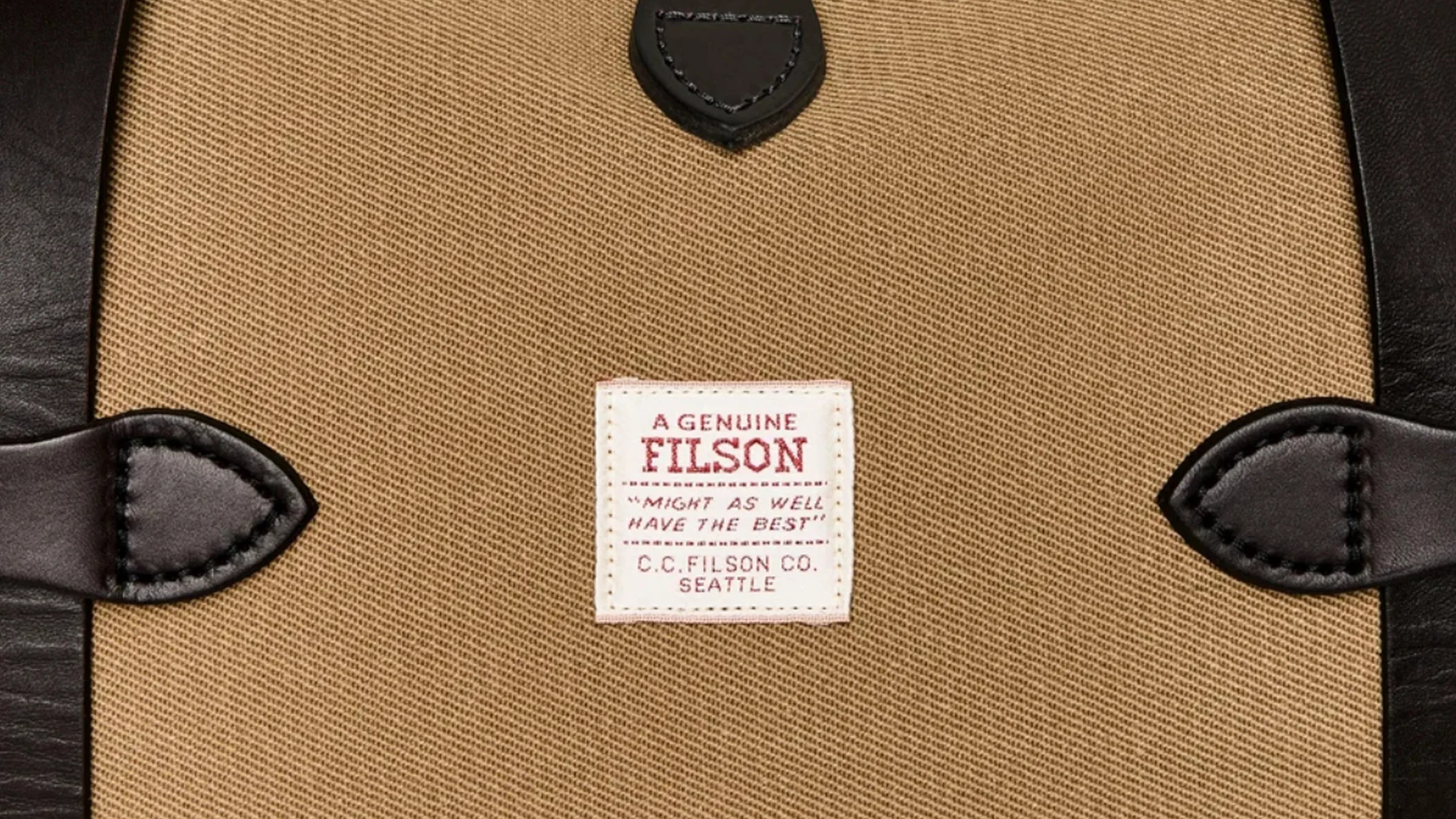 Close-up of a tan fabric bag with black leather trim and a white label reading "A Genuine Filson" and "C.C. Filson Co. Seattle.