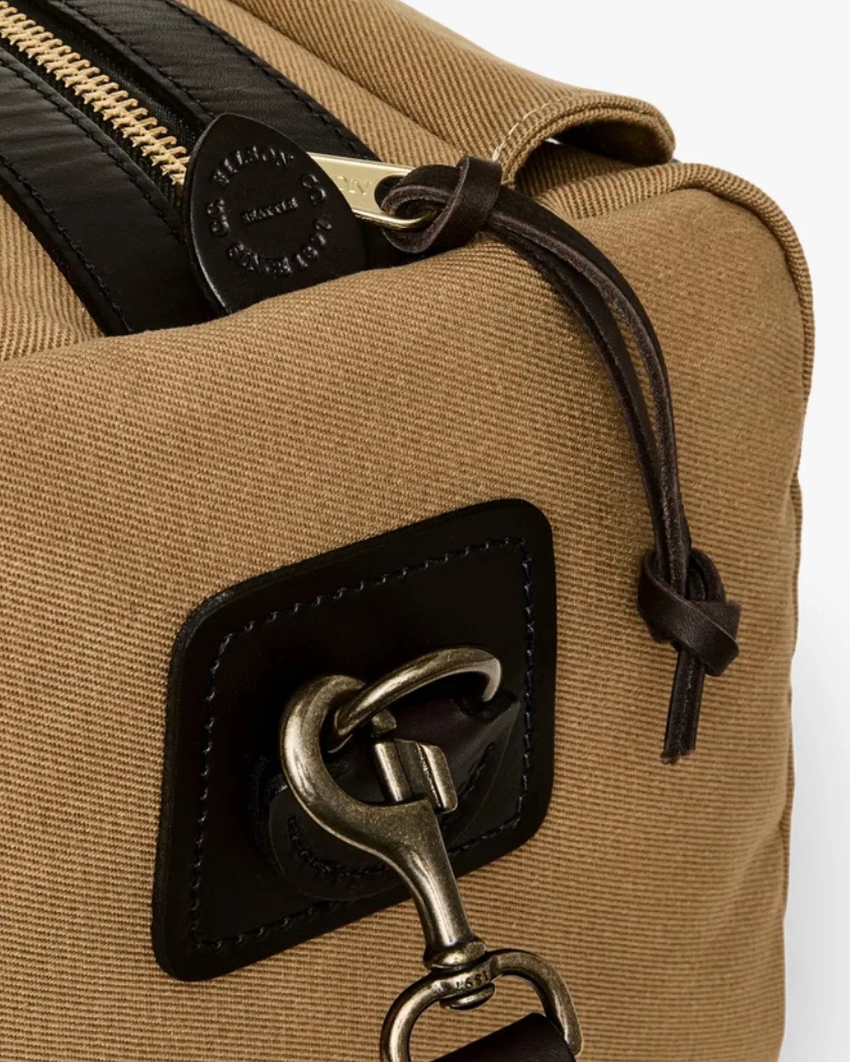 Close-up of tan canvas bag corner with black leather trim, brass zipper, and metal clasp attached to leather loop.