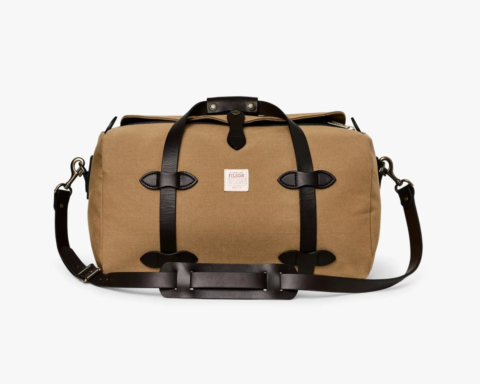 Tan canvas duffel bag with black leather straps and shoulder pad, featuring a small white label on the front.