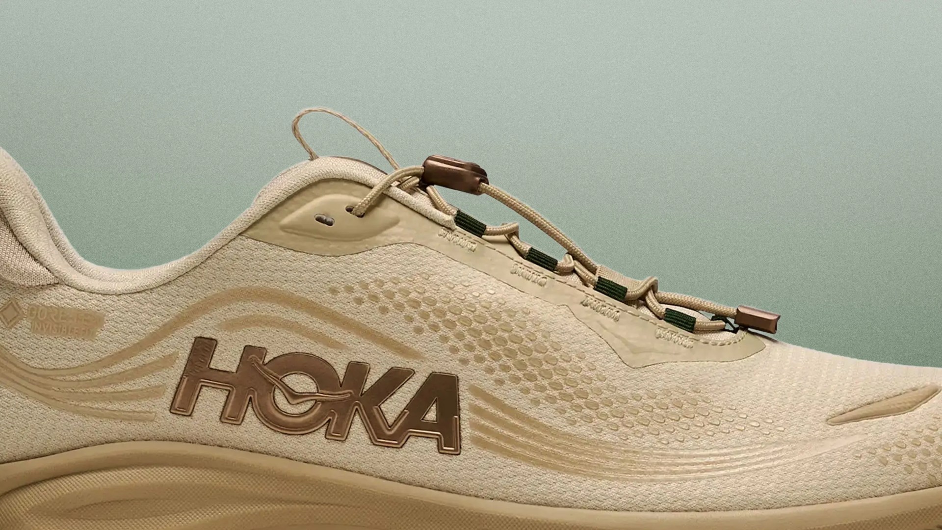 Beige Hoka running shoe with brown logo and toggle lace system against a muted green background.