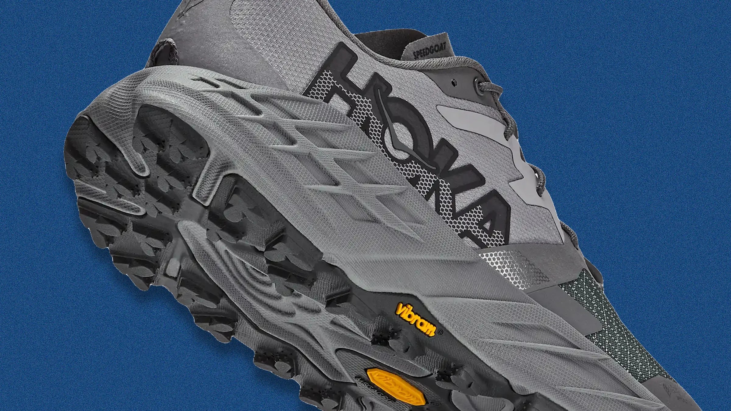 Gray HOKA trail running shoe with Vibram sole and black tread on a blue background.