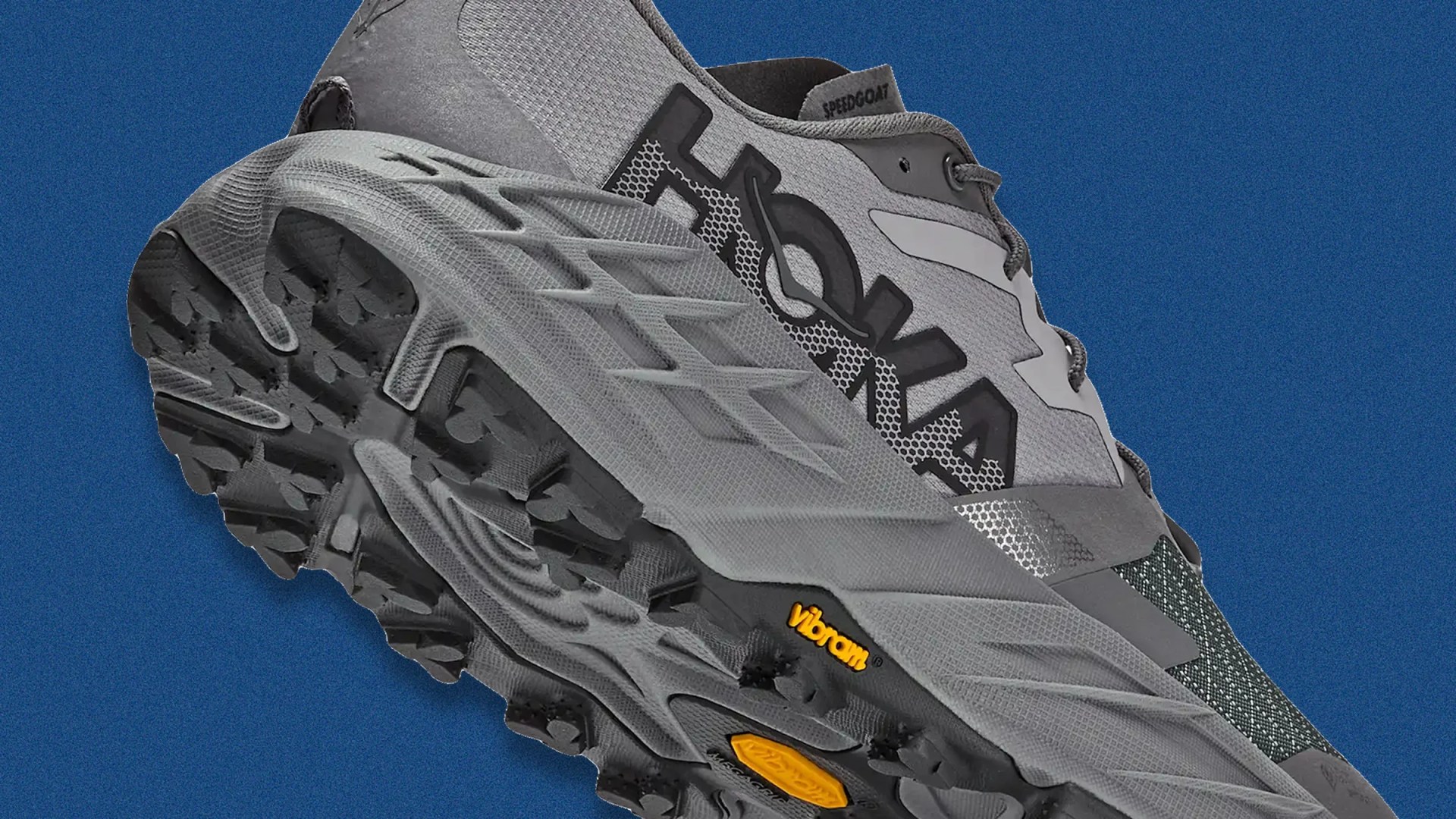 Gray HOKA trail running shoe with Vibram sole and black tread on a blue background.