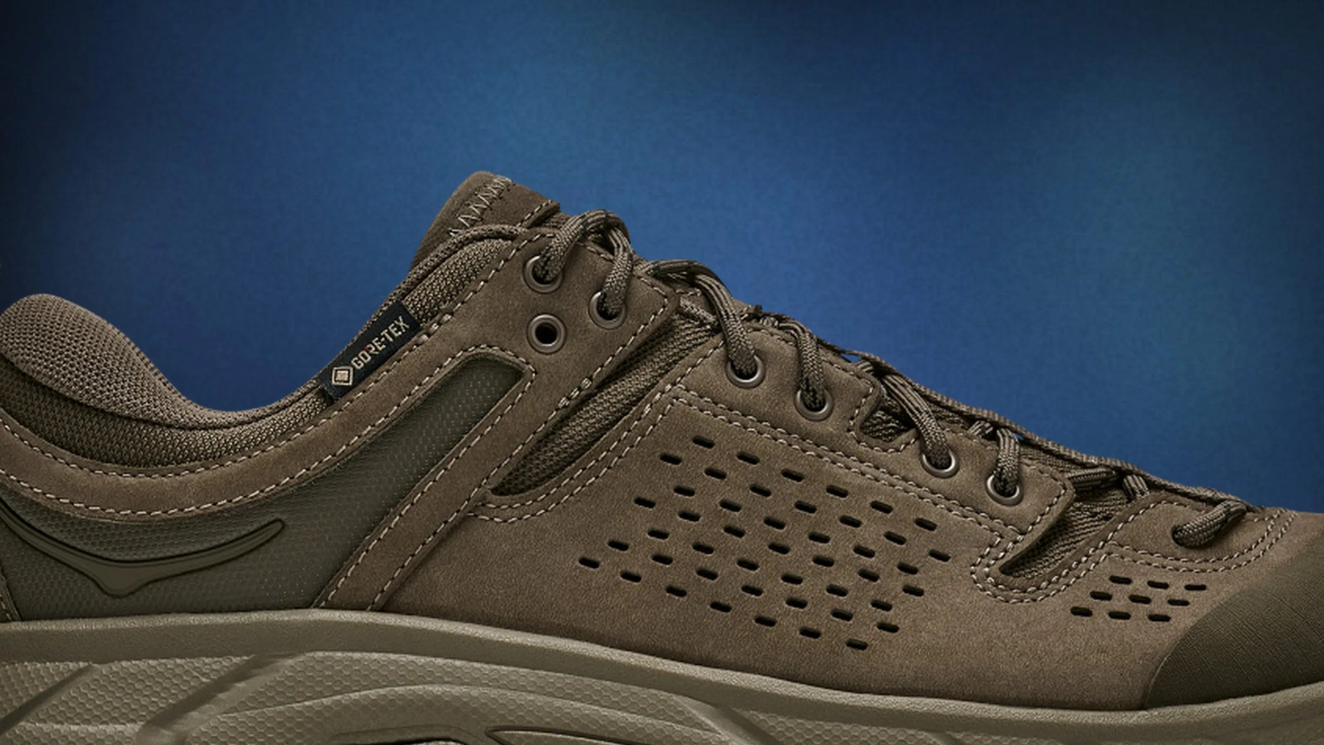 Brown Gore-Tex sneaker with perforated suede upper and thick textured sole against a blue background.