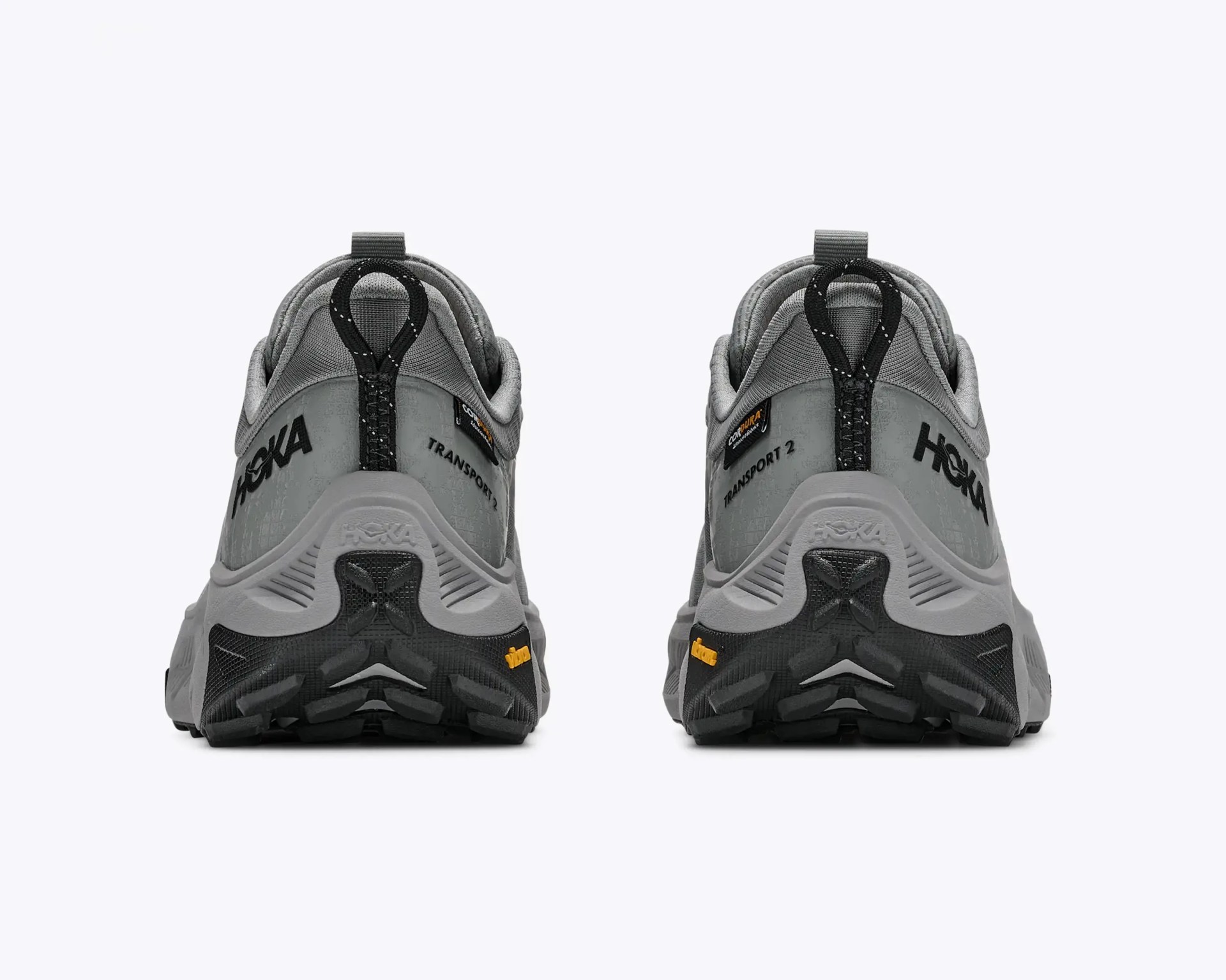 Pair of gray Hoka Transport 2 sneakers viewed from the back showing black and yellow Vibram soles.
