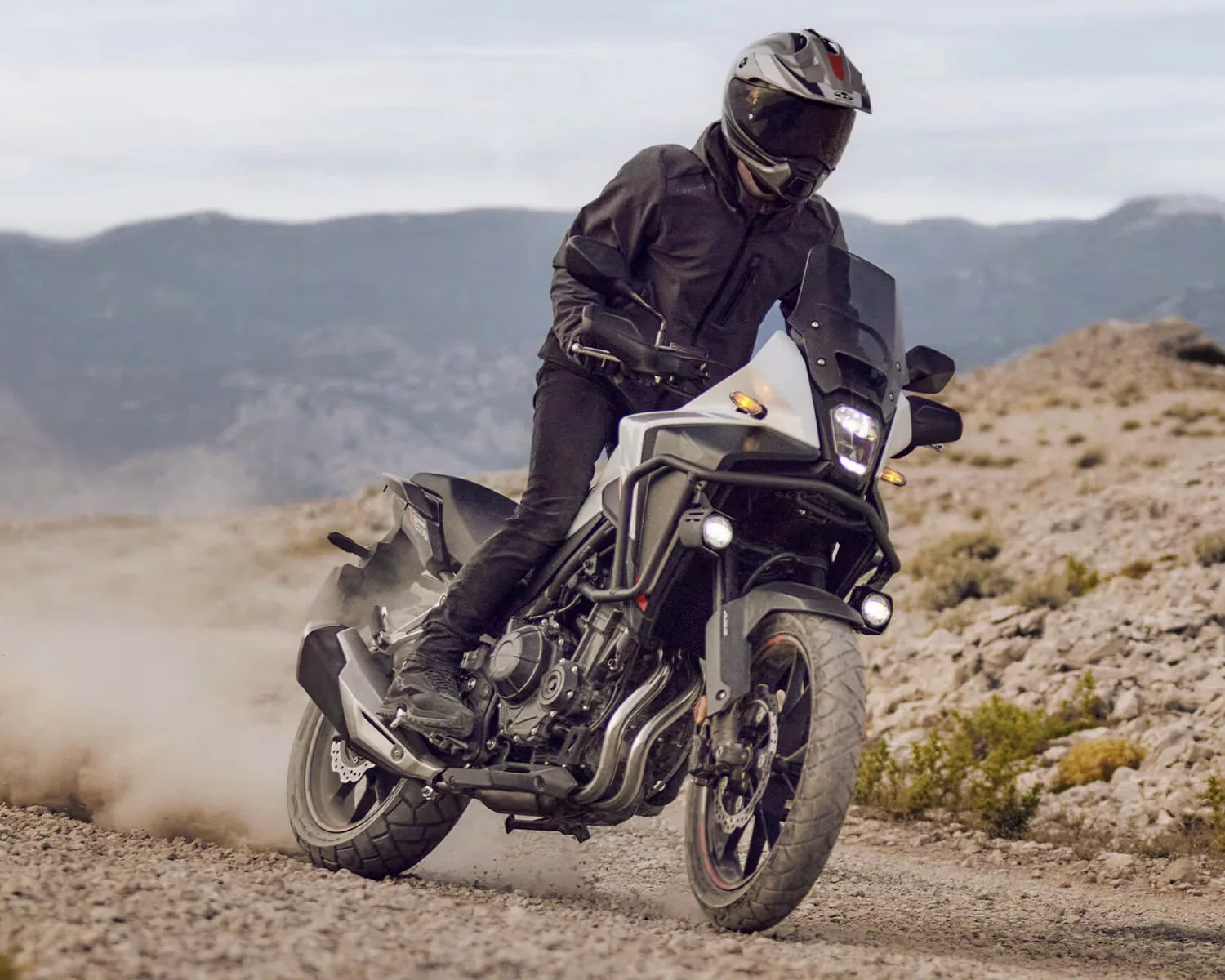 Motorcyclist in black gear riding a white adventure motorcycle on a rocky dirt trail with mountains in the background.