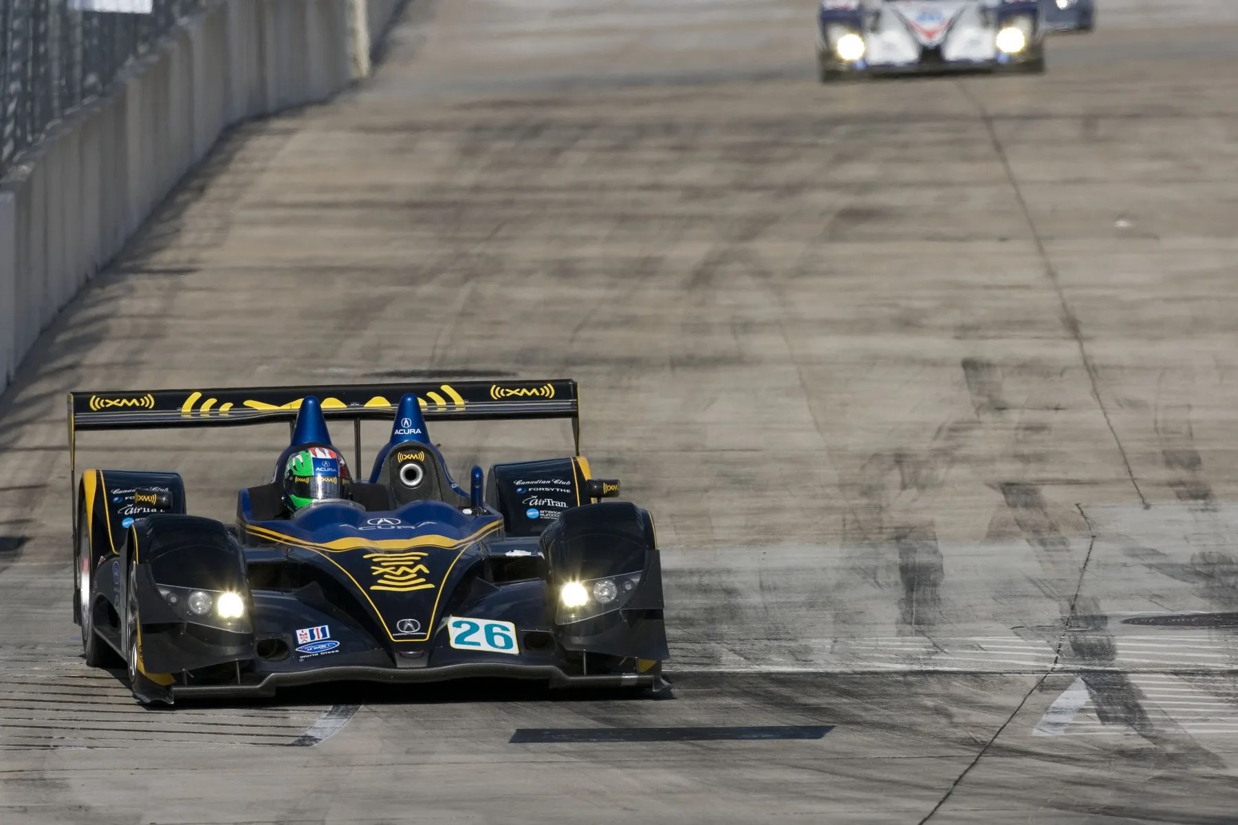Black and yellow Acura race car number 26 on a concrete race track with headlights on.