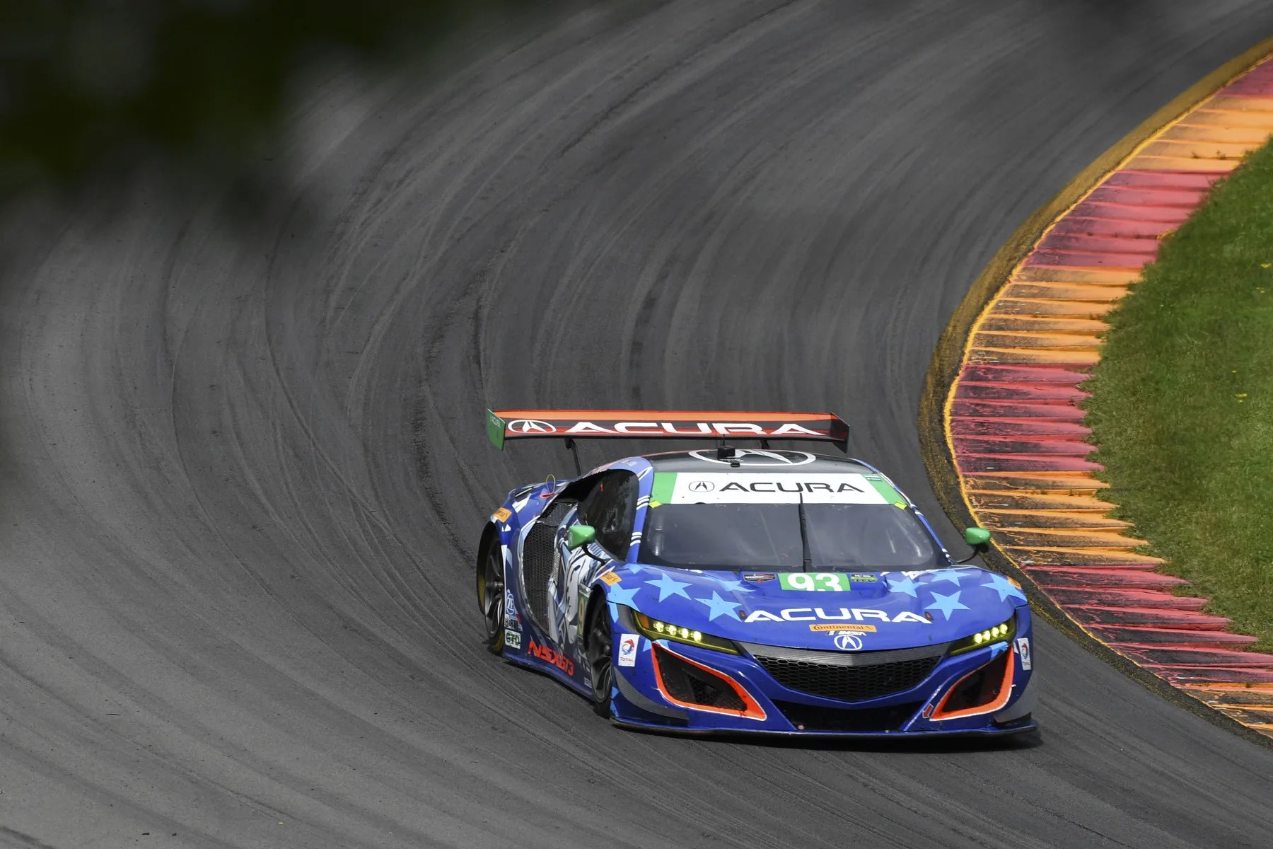 Blue Acura race car with stars and number 93 on a curved racetrack near red and yellow curbs.