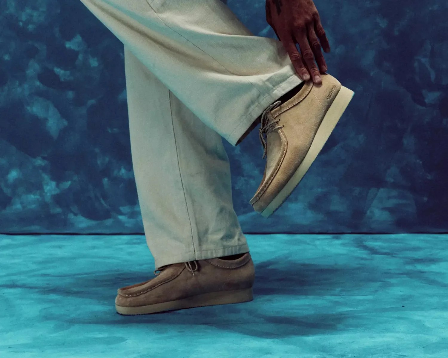 Beige suede Wallabee shoes with crepe soles worn with light-colored pants against a blue textured background.