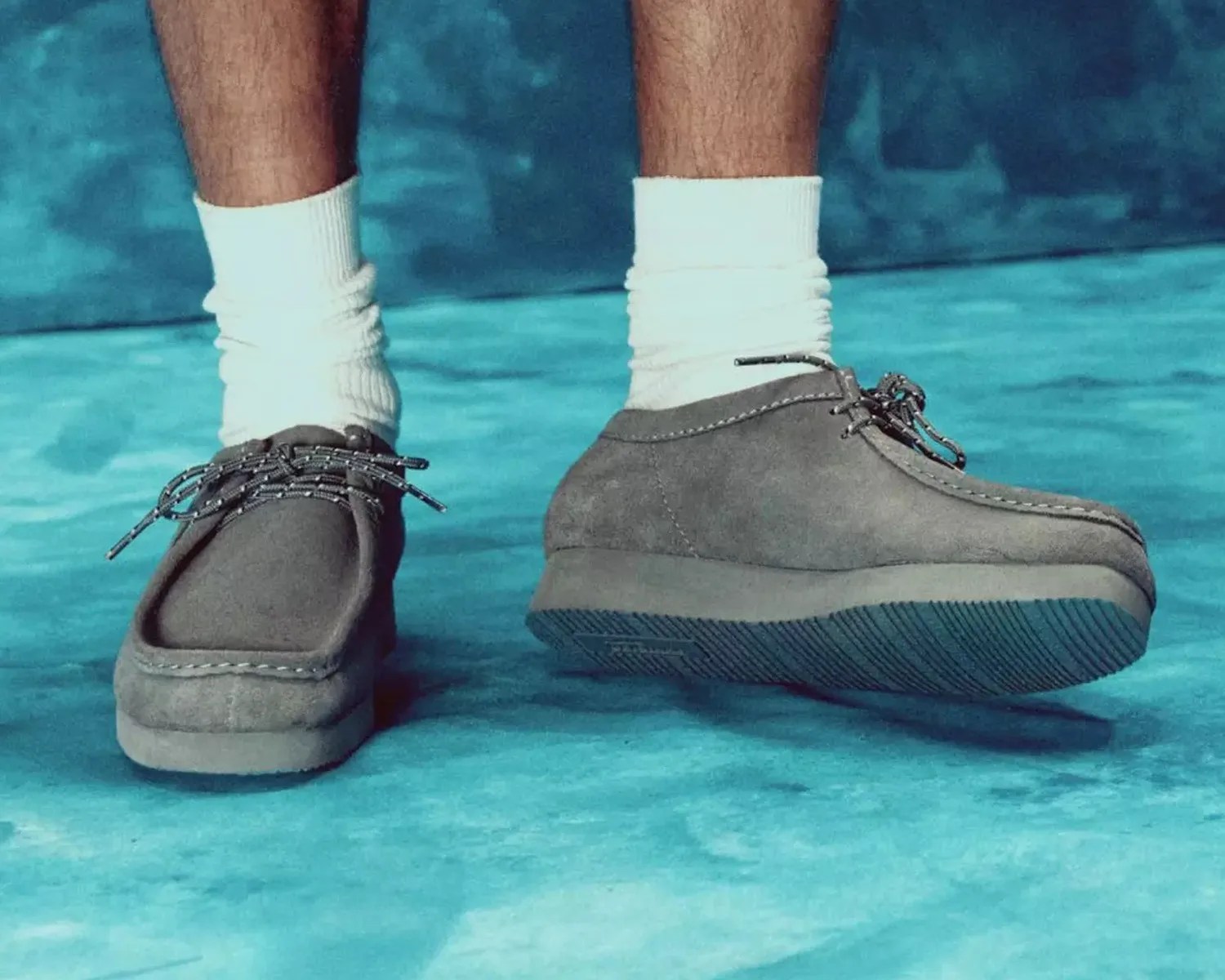 Gray suede moccasin-style shoes with black and white laces worn with white socks on a blue surface.