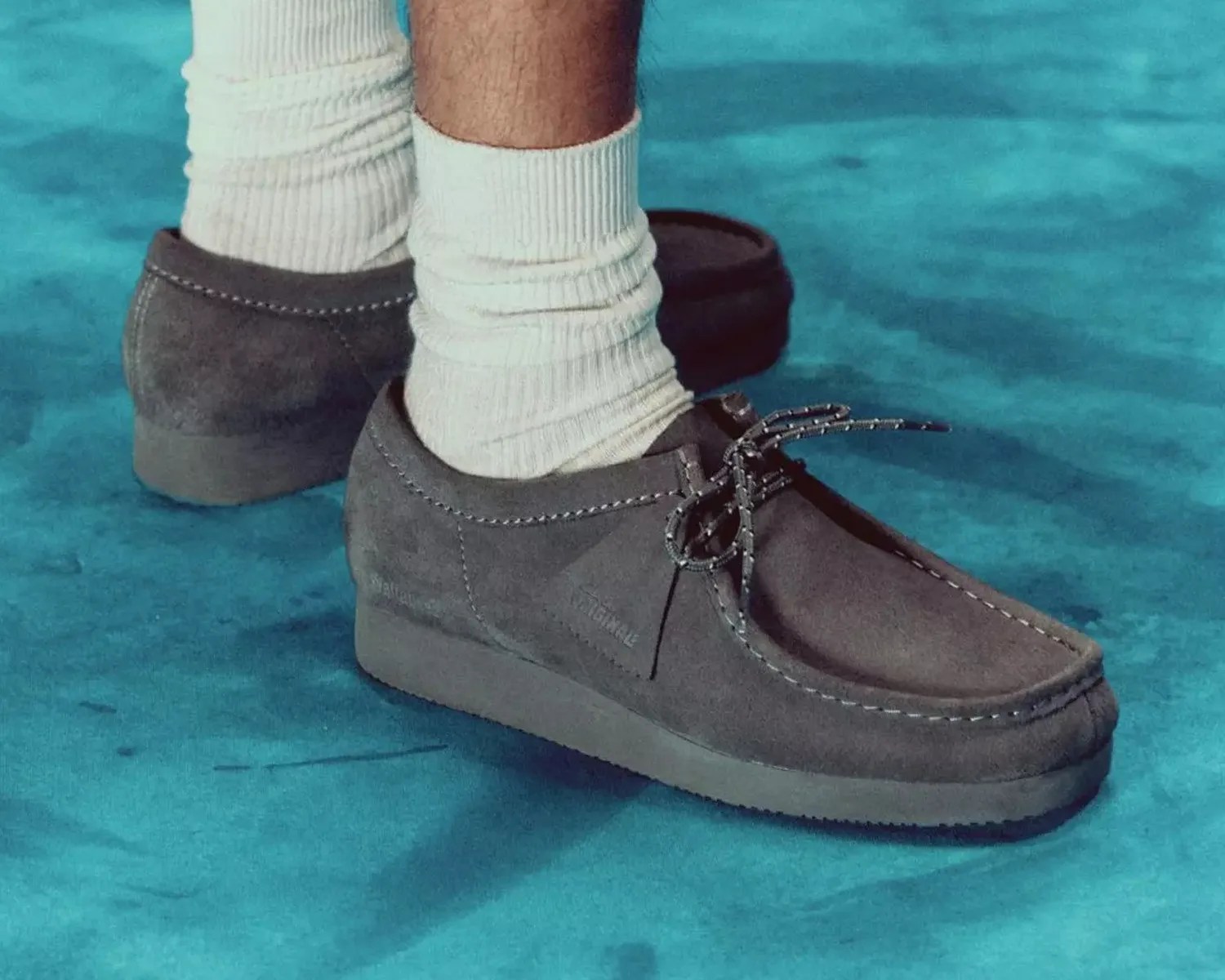 Gray suede moccasin-style shoes with black and white laces worn with white ribbed socks on a blue surface.
