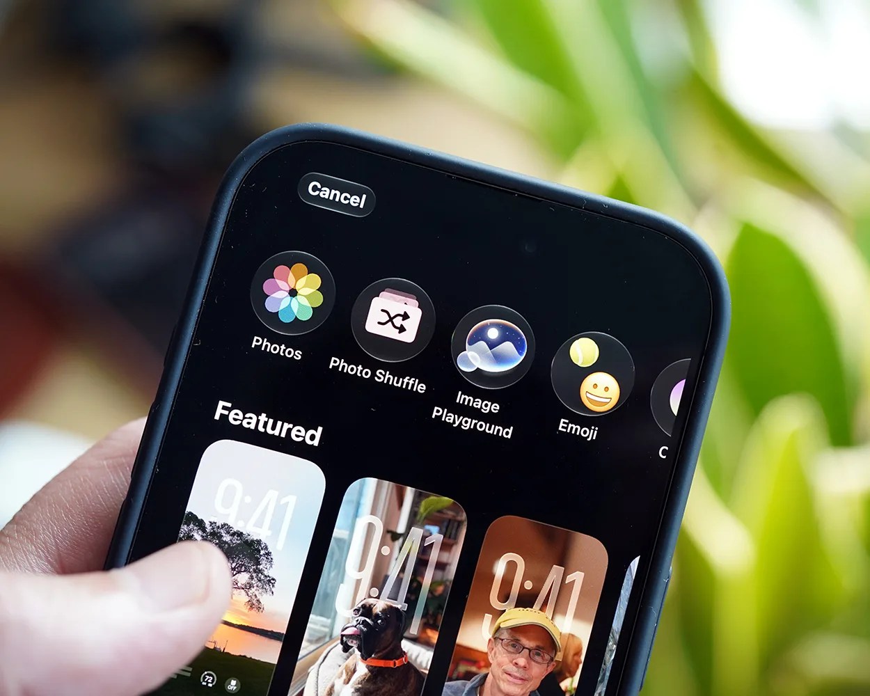 Close-up of a hand holding a smartphone displaying wallpaper customization options with icons for Photos, Photo Shuffle, Image Playground, and Emoji.