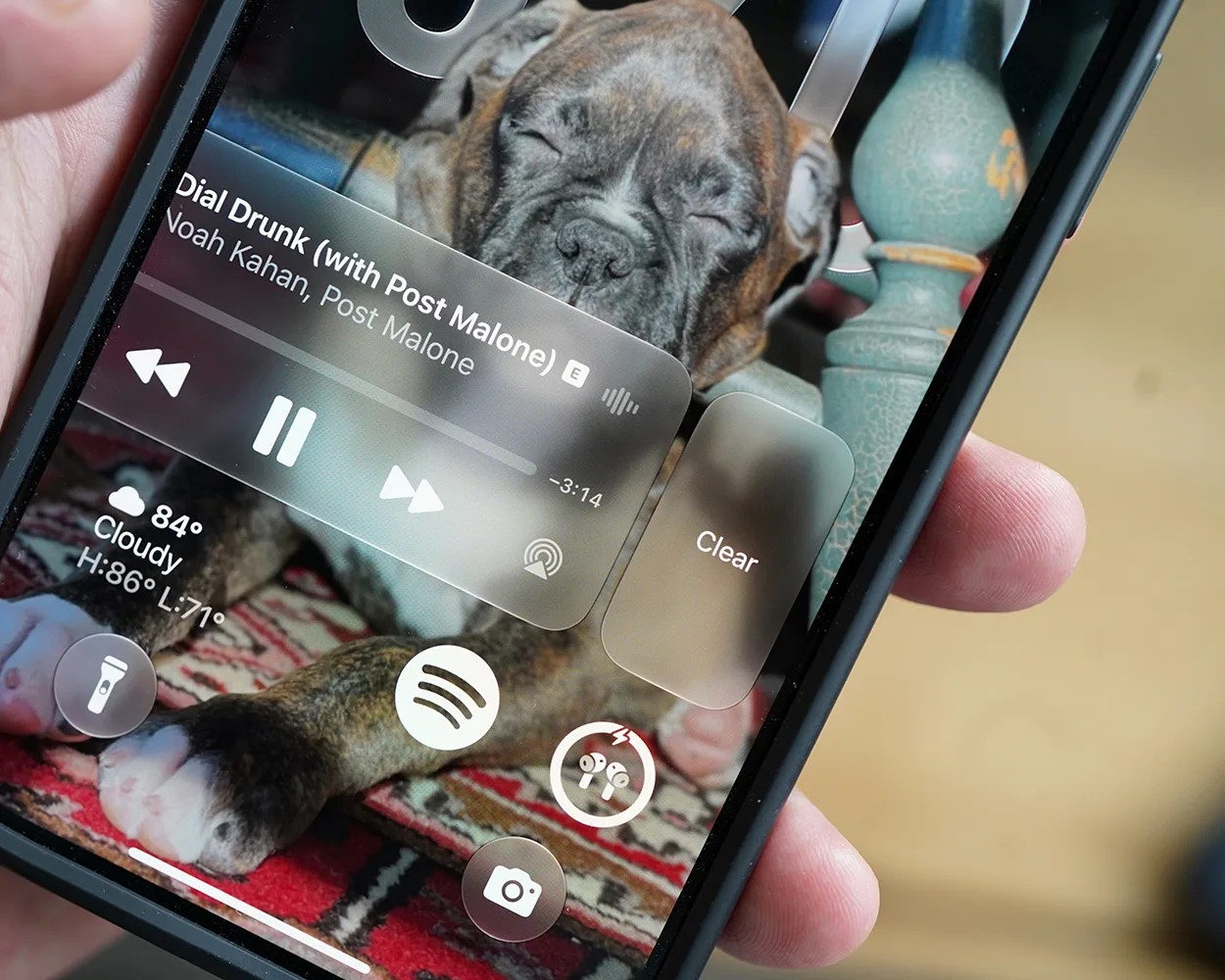 Smartphone screen showing music player with song "Drunk (with Post Malone)" by Noah Kahan, weather info, and a background image of a sleeping brindle puppy on a patterned rug.