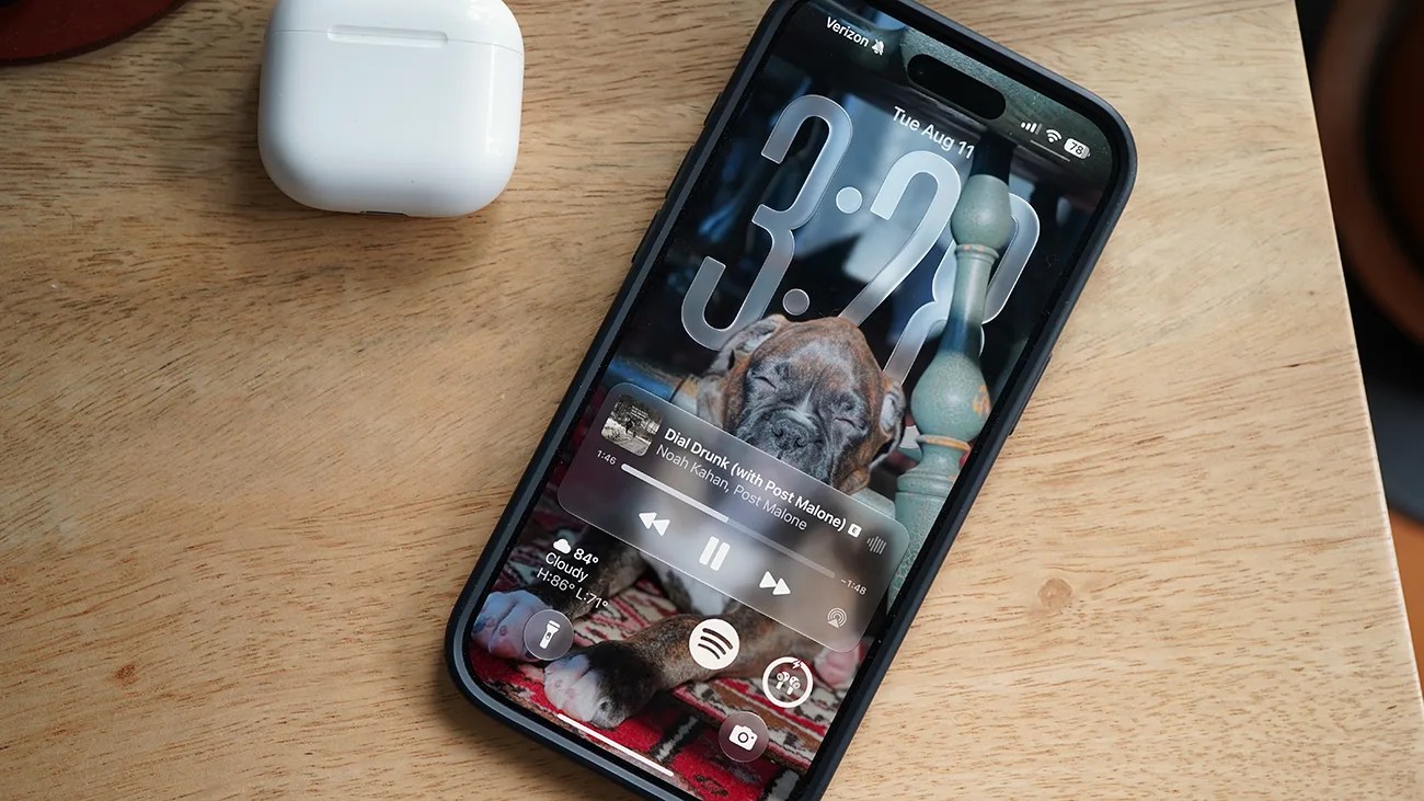 Smartphone with a black case displaying a music player screen and a puppy wallpaper, next to a white wireless earbud case on a wooden surface.