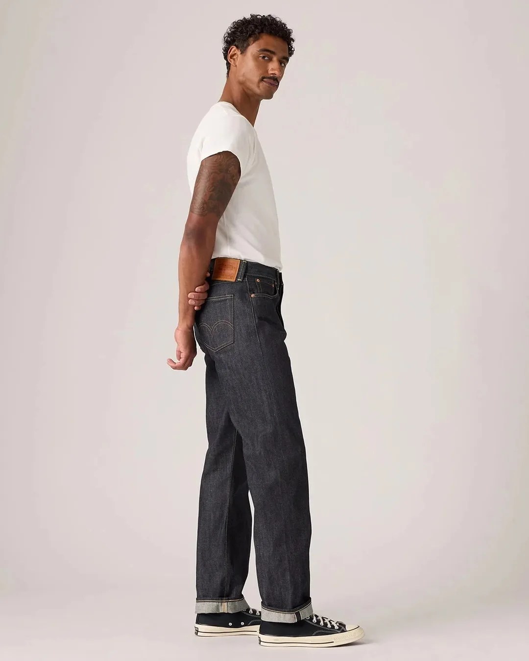Man wearing dark raw denim jeans with cuffs, a white t-shirt, and black low-top sneakers standing sideways.