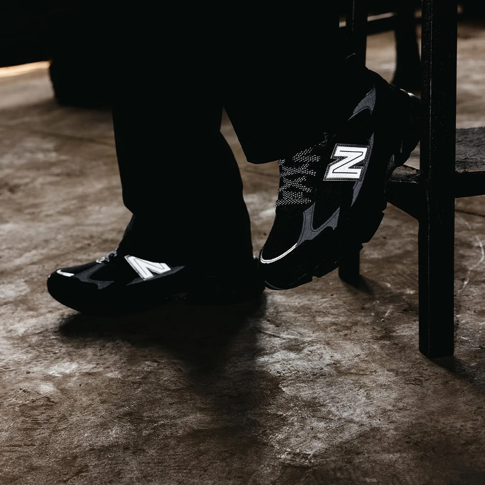 Black New Balance sneakers with white "N" logo worn by a person sitting on a chair on a concrete floor.