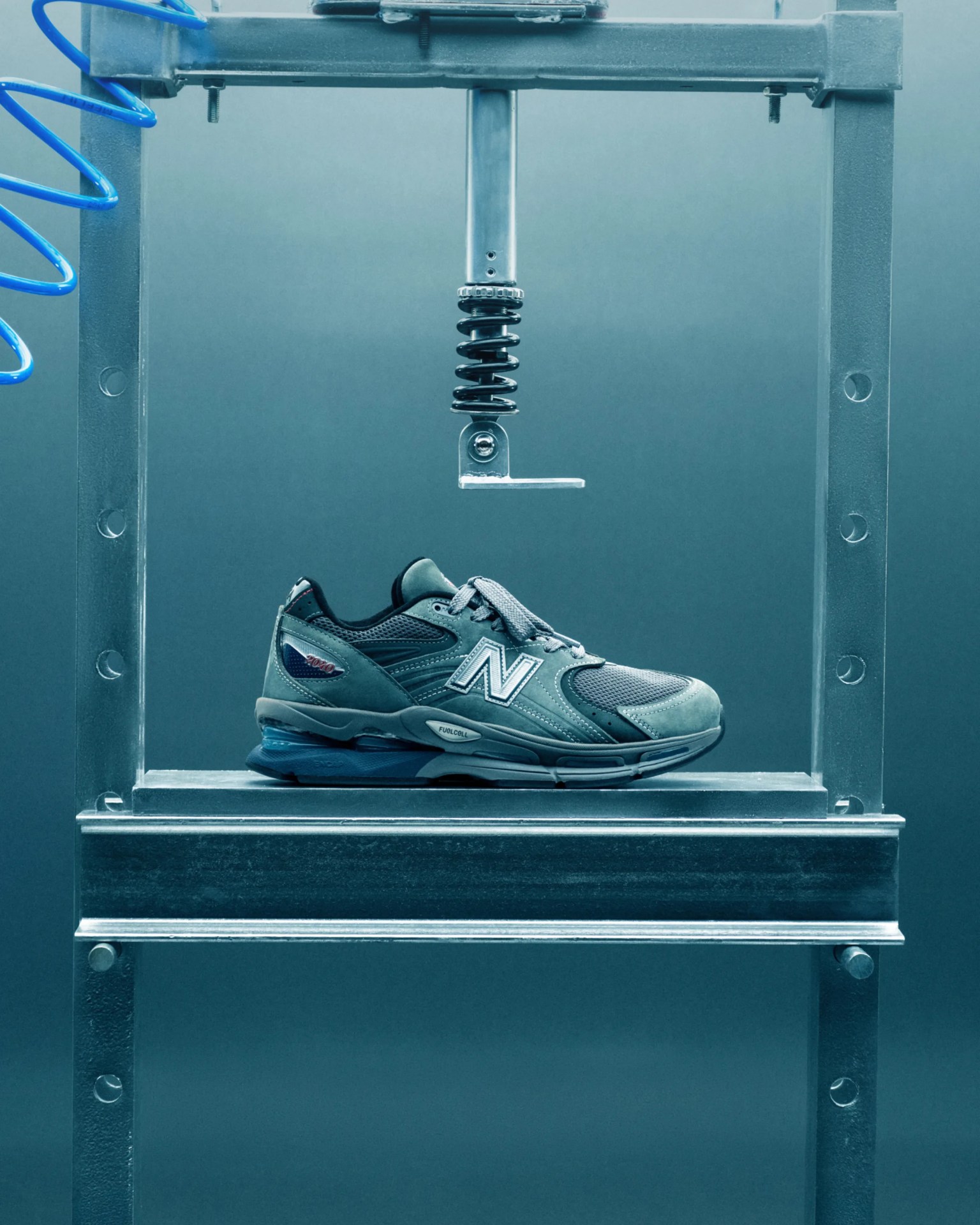 Gray New Balance sneaker with blue accents under a metal compression testing machine.