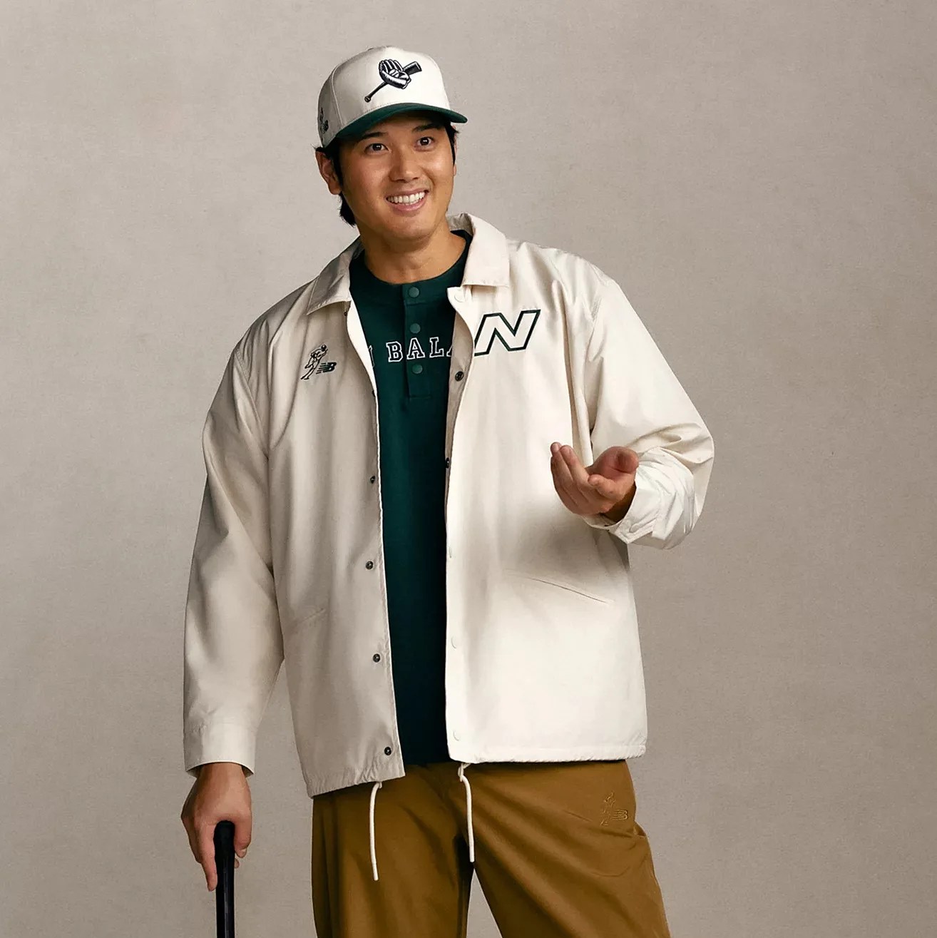 Man wearing a cream button-up jacket with logos, green shirt, brown pants, and a white baseball cap holding a black cane.