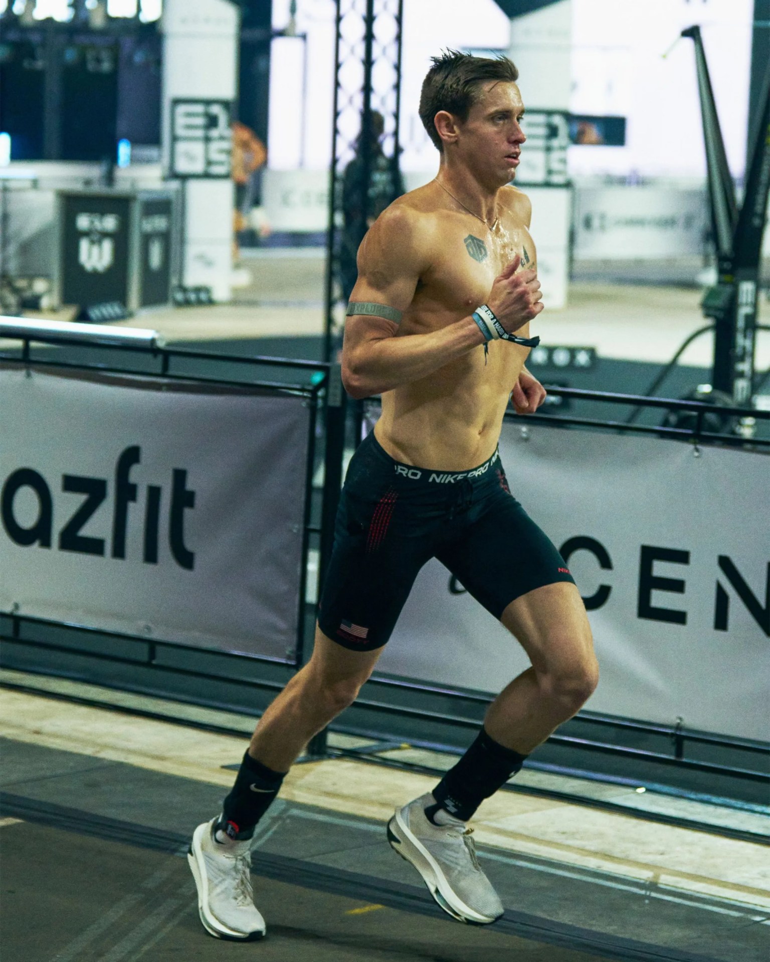 Shirtless male runner wearing black Nike Pro shorts, black socks, and white running shoes indoors.