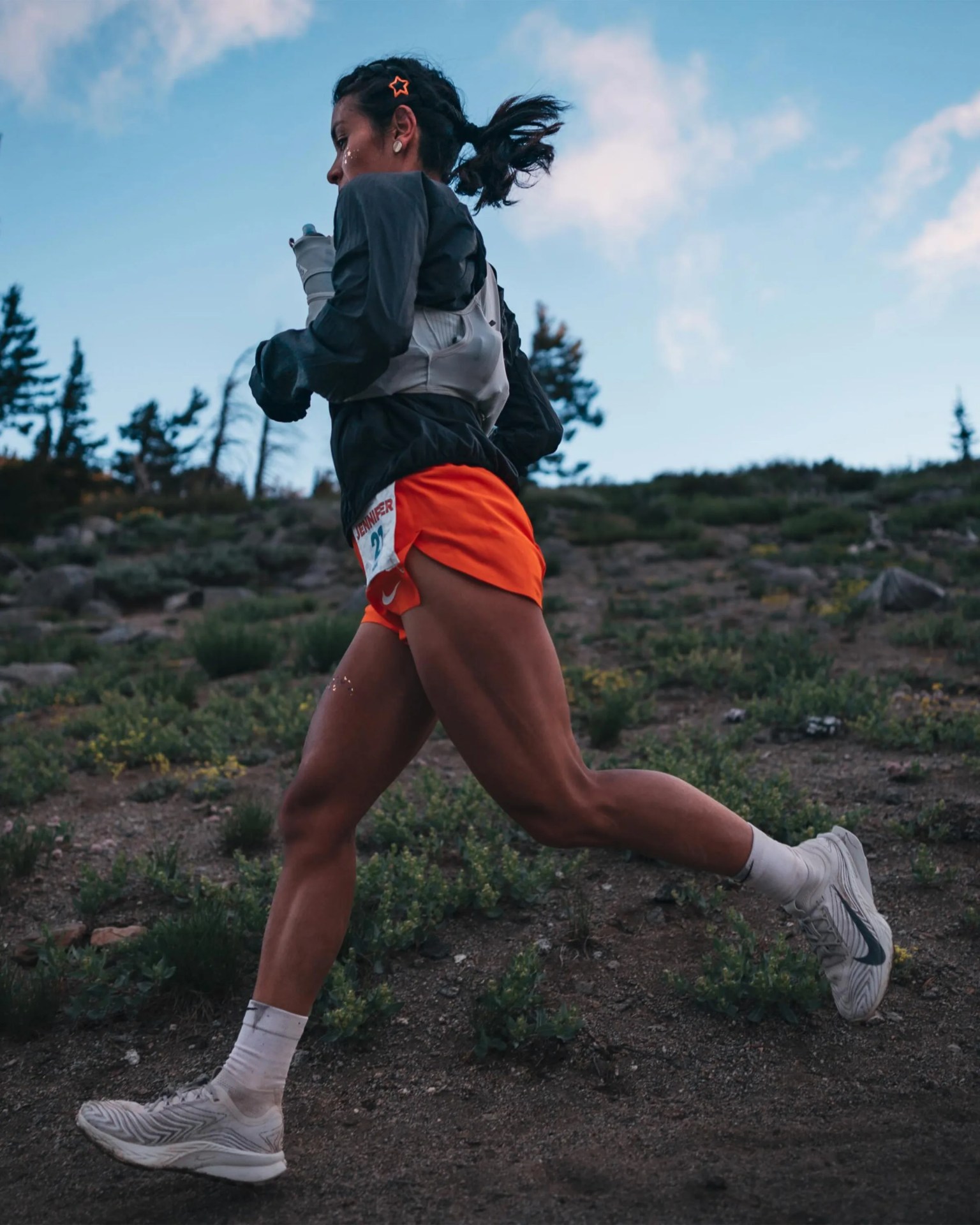 Runner wearing orange shorts, black jacket, and white Nike shoes running on a trail with greenery and trees in the background.