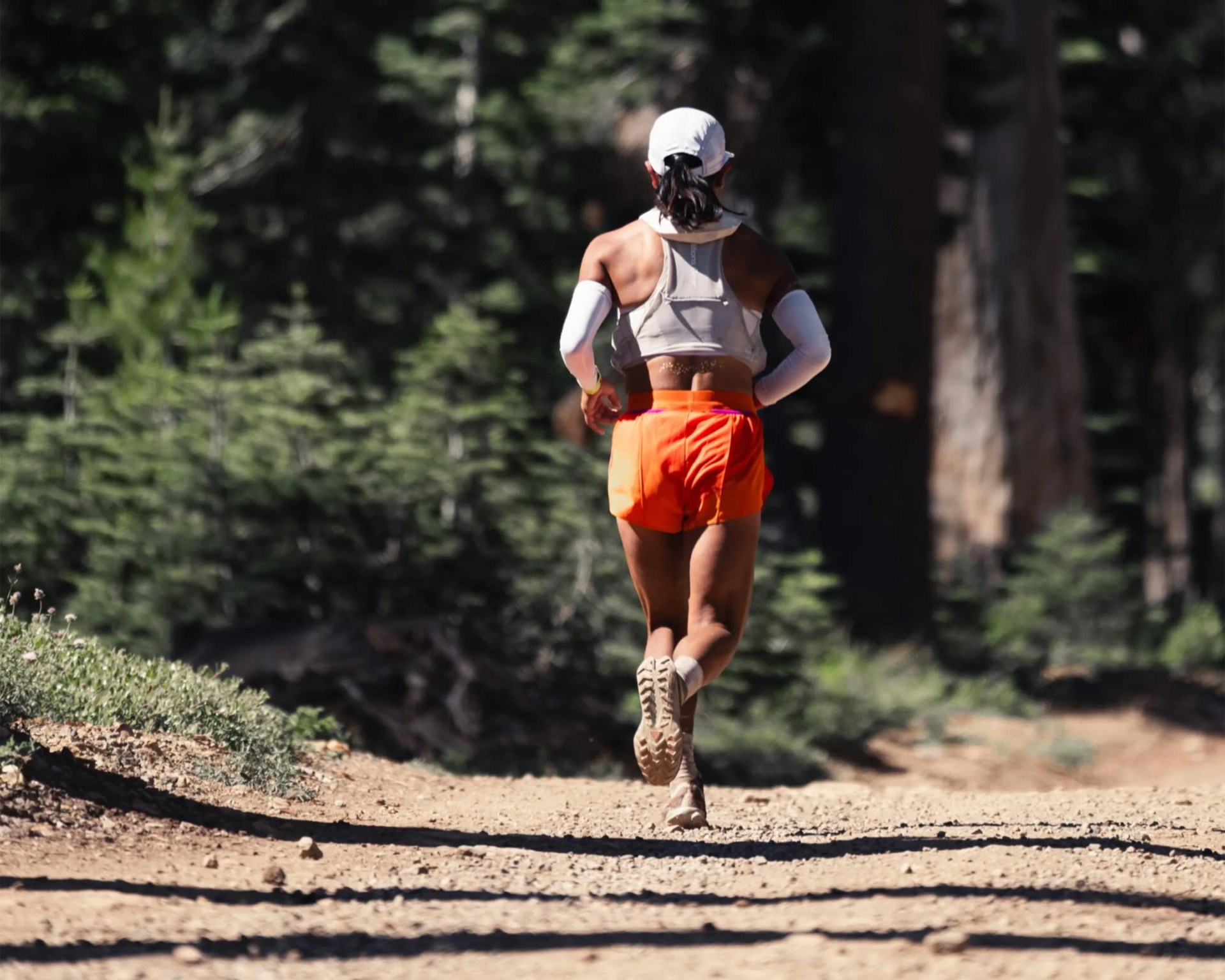 Runner in orange shorts and white sleeveless top running on a dirt trail surrounded by trees.